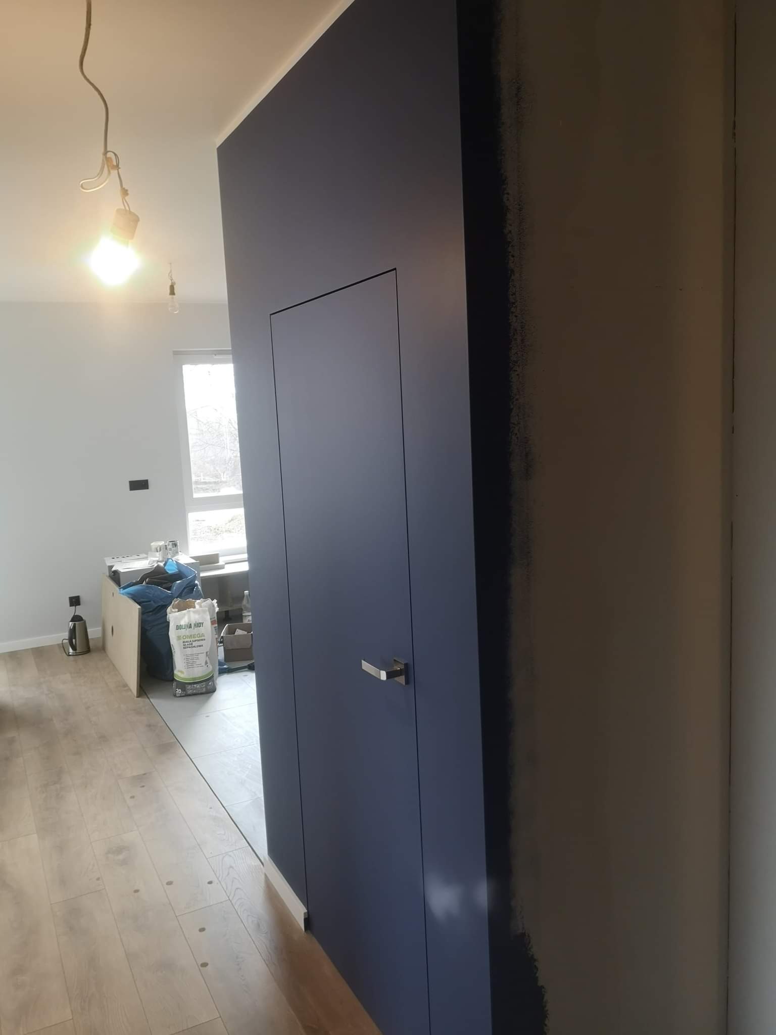 Niebieskie drzwi ukryte w ścianie, widoczny worek z gładzią i surowe wykończenie ściany w tle, lampa na kablu.