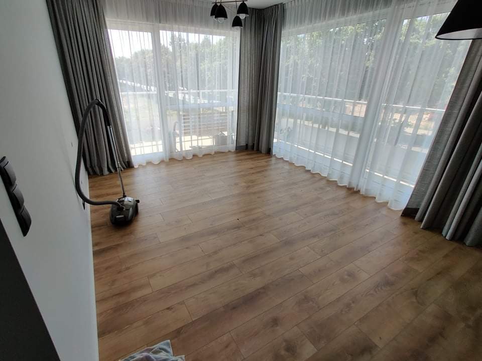 Jasne wnętrze z parkietem w odcieniach brązu, widok na balkon przez okna z firanami i zasłonami, odkurzacz przy ścianie.