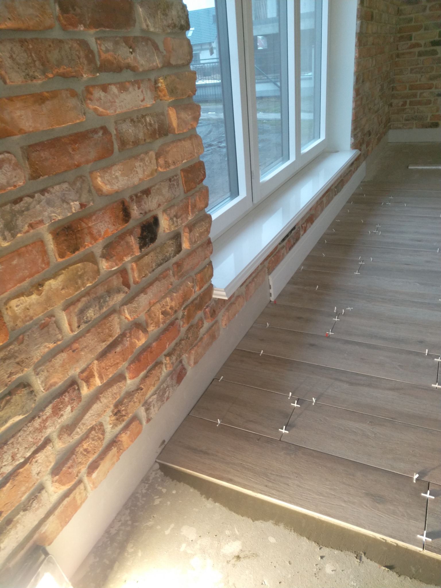 Fragment wnętrza w trakcie prac: ceglana ściana, białe okno, podłoga z paneli układana z użyciem krzyżyków dystansowych.