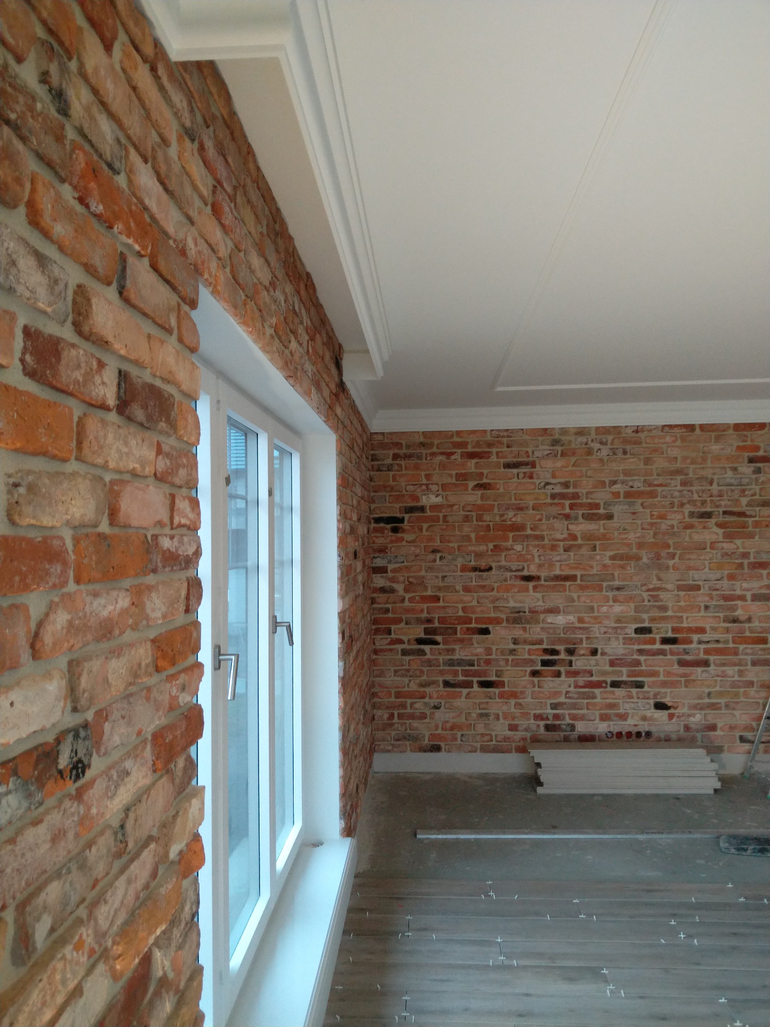 Ściana z cegły klinkierowej z białym oknem i listwami przysufitowymi; widoczne płyty gipsowe i poziomica na podłodze z przygotowanym układaniem płytek.