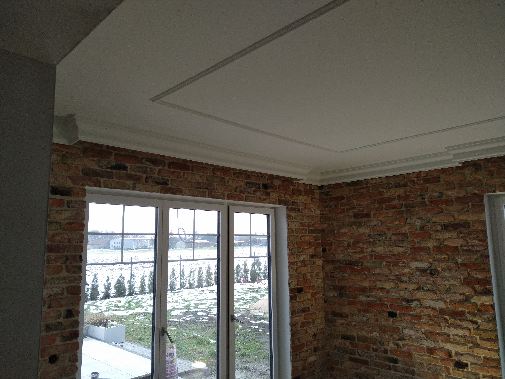 Ceglana ściana wewnętrzna z białymi, dwuskrzydłowymi oknami i ozdobnym wykończeniem sufitu z listwami.