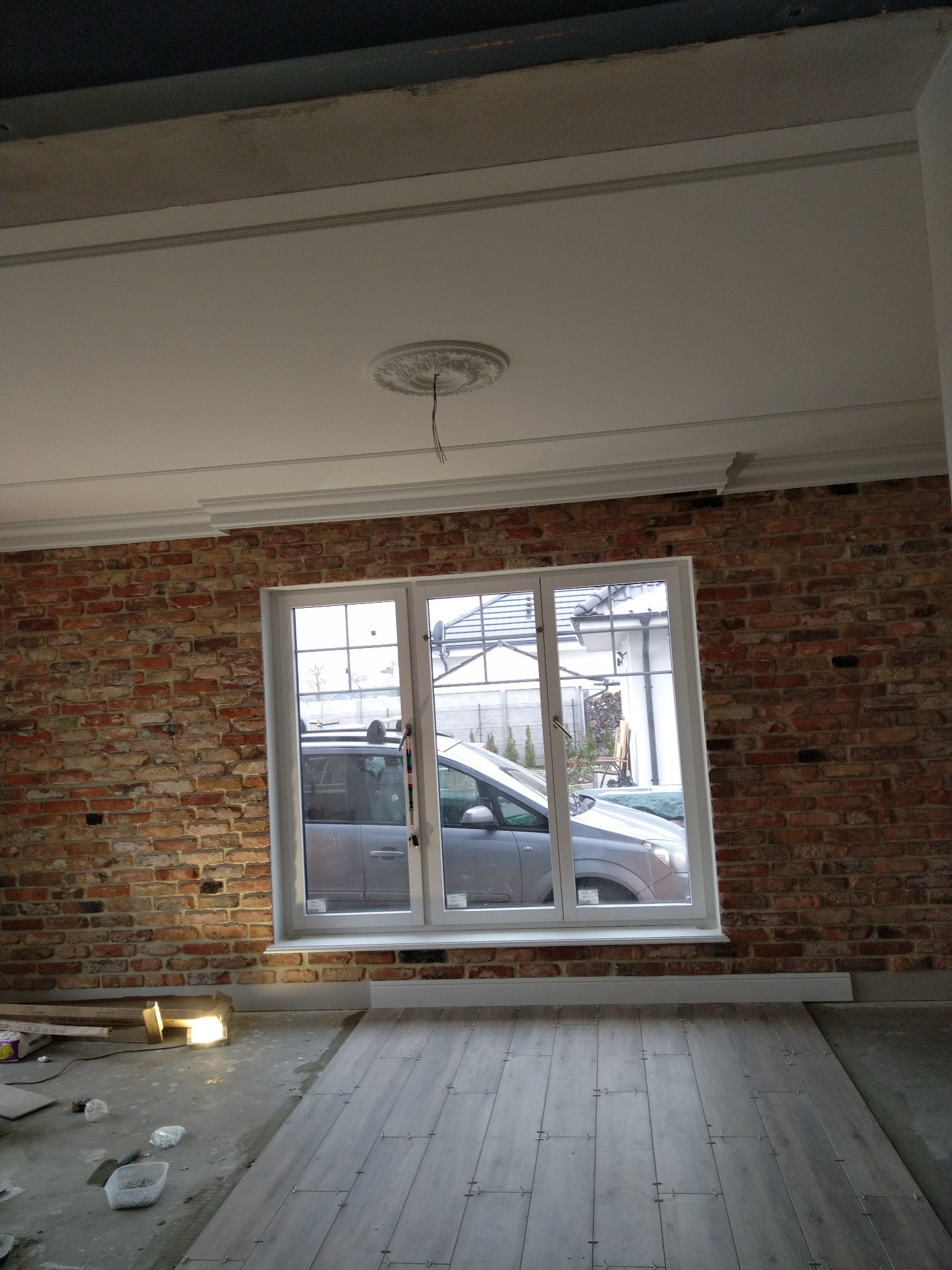 Pokój w trakcie remontu: fragment ściany z cegły, białe okno z widokiem na samochód, ułożona część szarych paneli podłogowych, widoczne listwy sufitowe i dekoracyjny element na suficie...