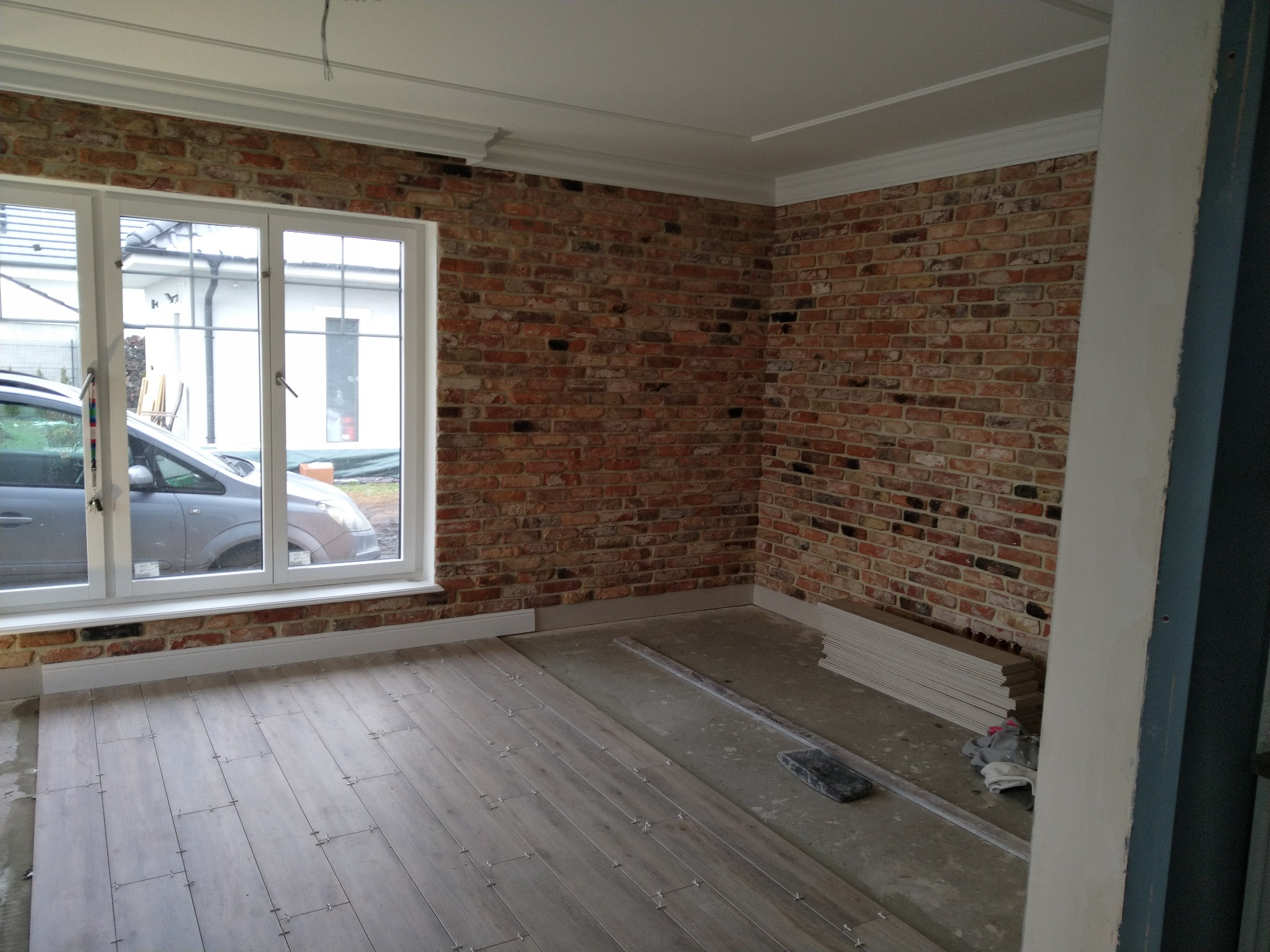 Układanie paneli podłogowych w pomieszczeniu z ceglaną ścianą i białymi listwami przypodłogowymi, widoczne krzyżyki dystansowe i stos paneli w rogu.