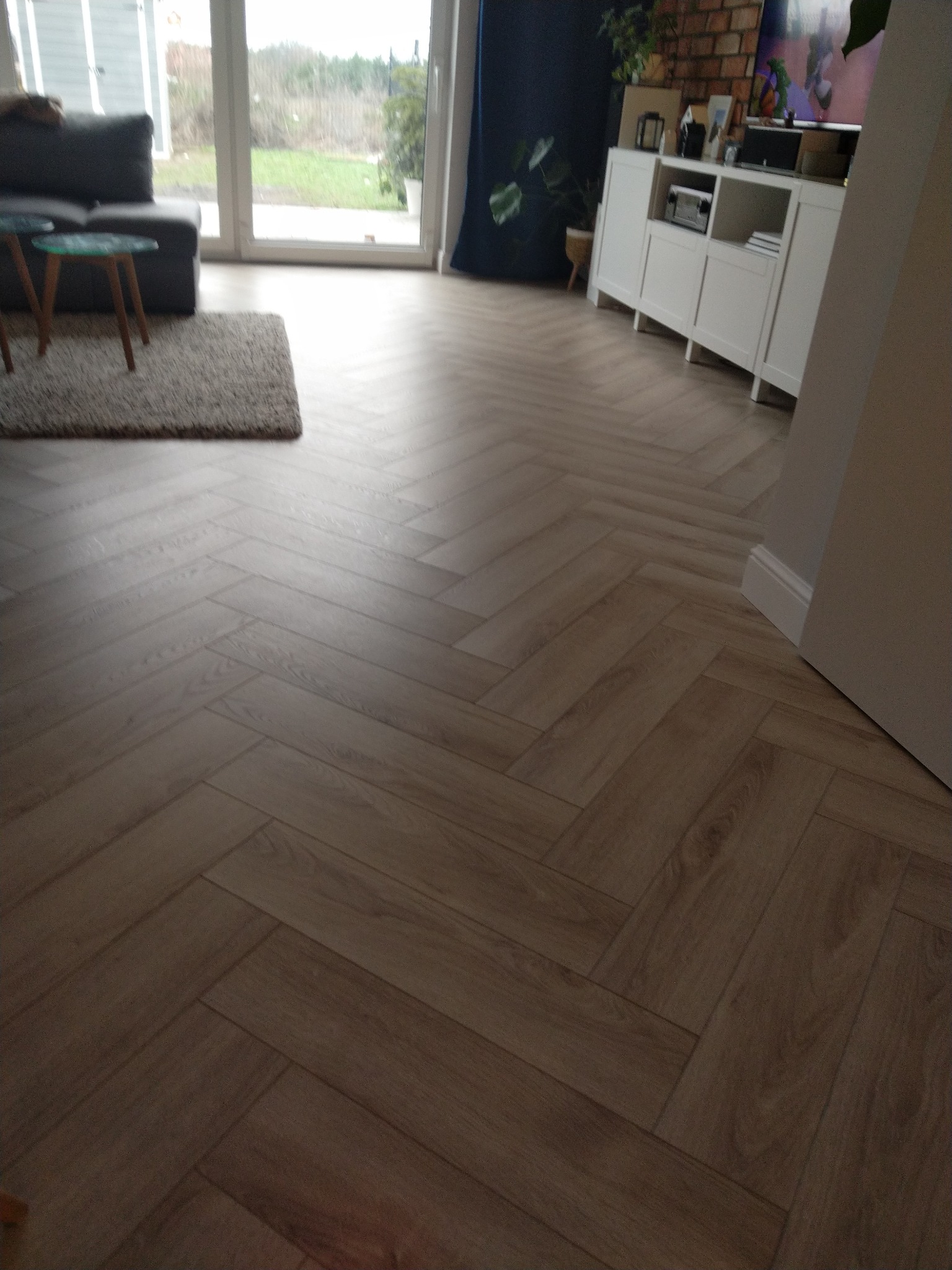 Parkiet ułożony w jodełkę francuską, widok na salon z sofą, stolikami i szafką RTV, domknięty widokiem na ogród przez przeszklone drzwi tarasowe.