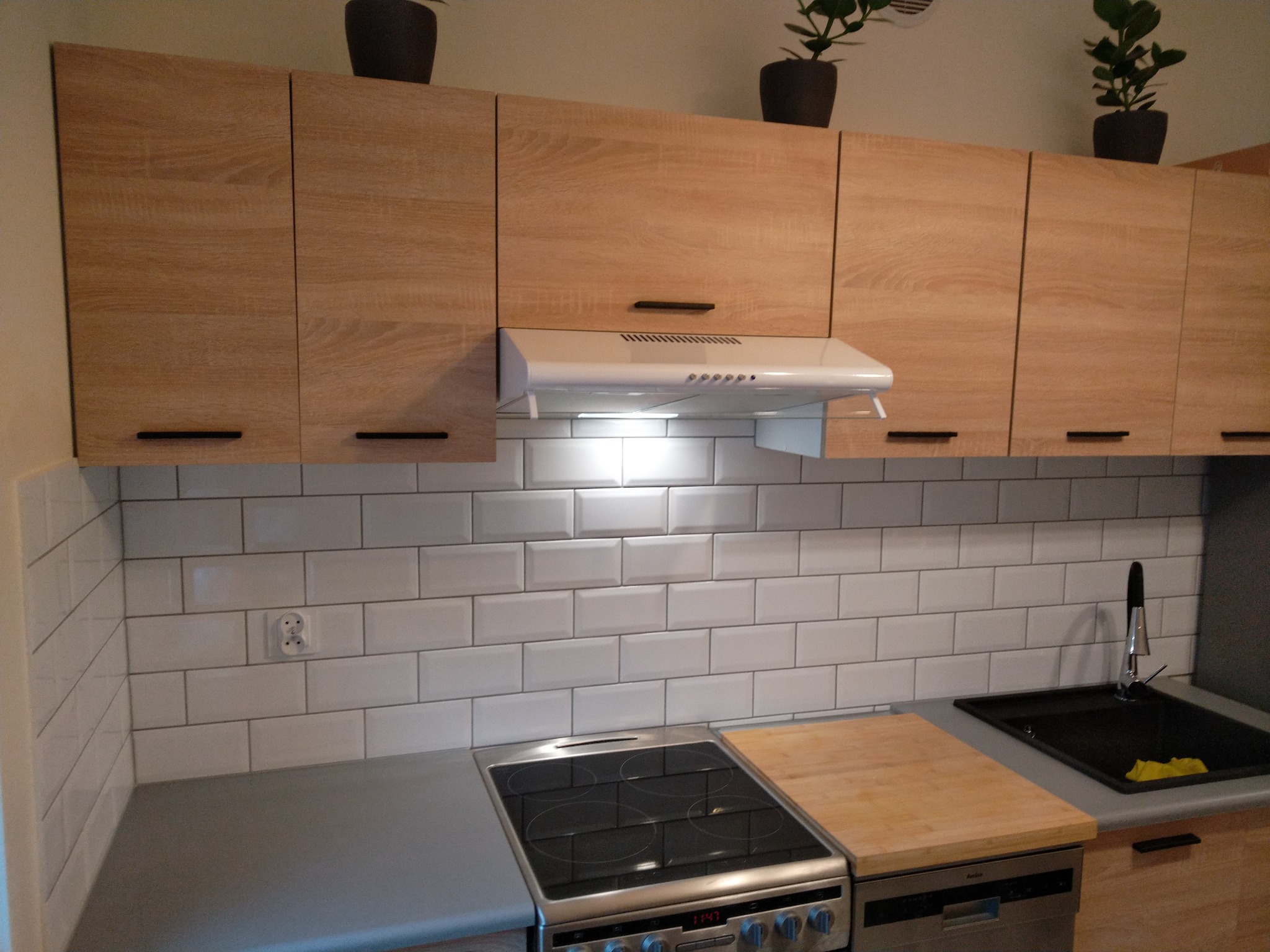 Jasna kuchnia z drewnianymi szafkami, białymi płytkami metra, czarnym zlewem i roślinami doniczkowymi na górnych szafkach.