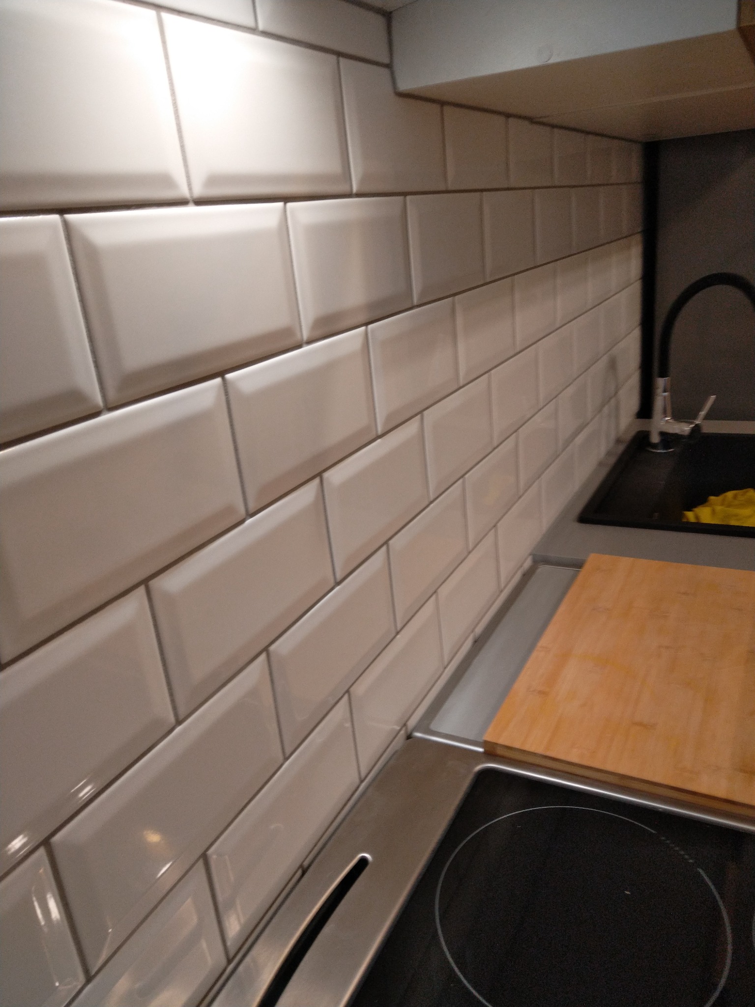 Białe, błyszczące płytki typu metro na ścianie w kuchni, widoczny fragment zlewu z czarną baterią oraz fragment płyty indukcyjnej z drewnianą deską do krojenia.