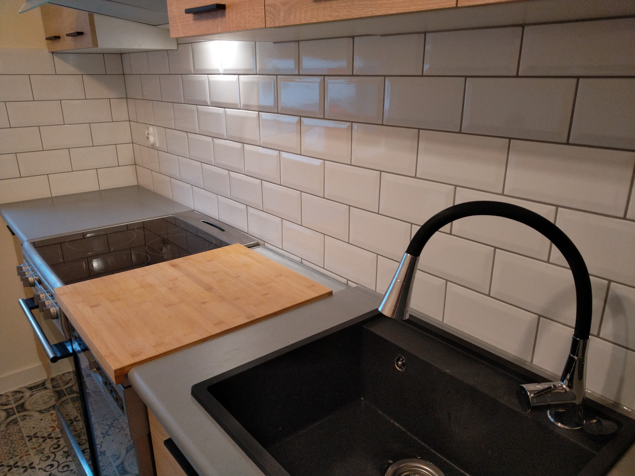 Nowoczesna kuchnia z białymi, cegiełkowymi płytkami, czarnym zlewem z elastyczną wylewką, drewnianą deską do krojenia i płytą indukcyjną.