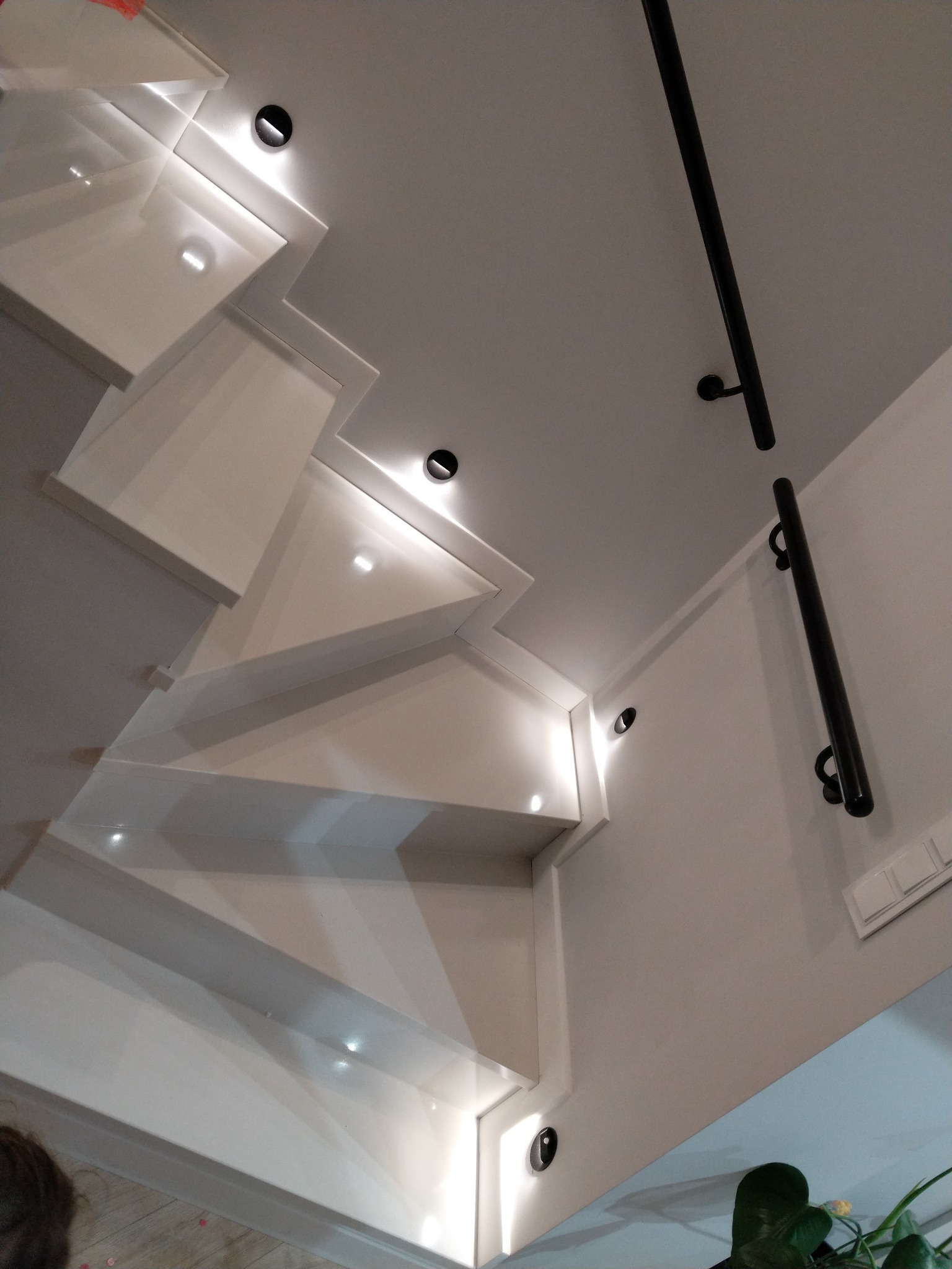 Nowoczesne, białe schody z podświetleniem punktowym i czarną, metalową poręczą zamontowaną na ścianie. Widok z dołu, podkreślający minimalistyczny design i oświetlenie.