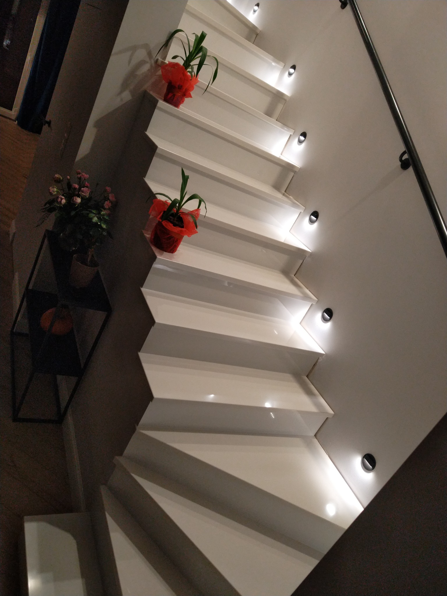 Białe schody z podświetleniem LED w ścianie, ozdobione doniczkami z roślinami, widok z dołu.