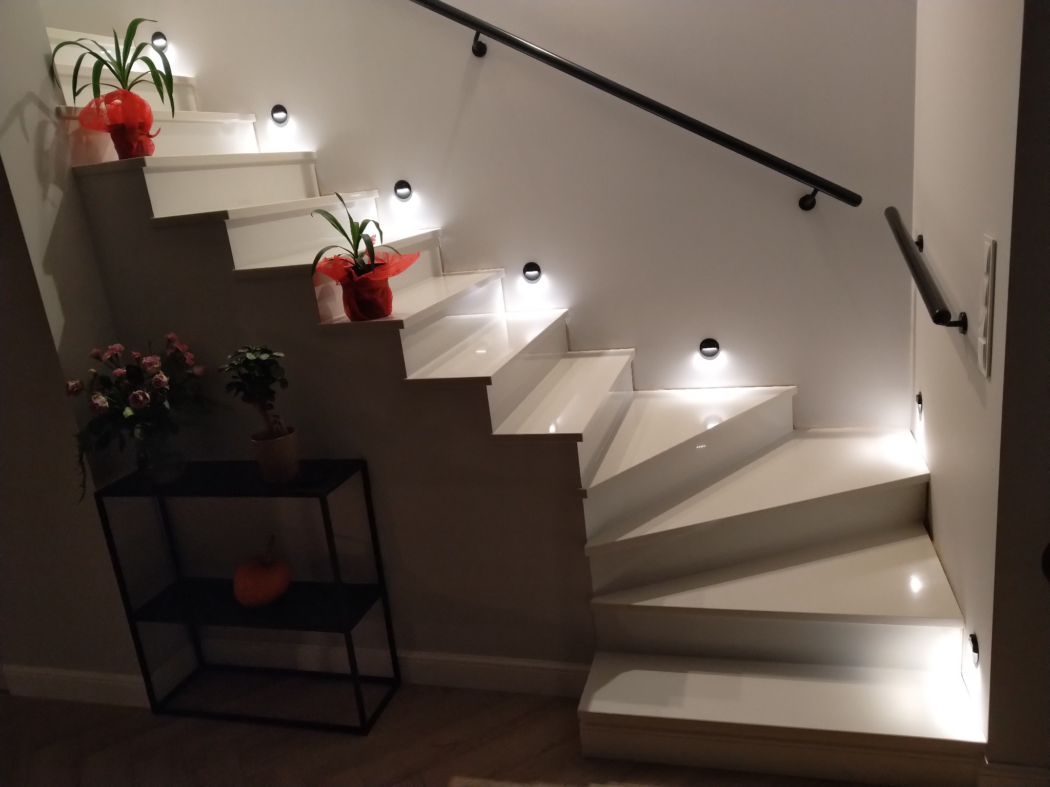 Białe schody z podświetleniem LED w ścianie, czarna poręcz, doniczki z kwiatami na stopniach i stolik z dekoracjami obok schodów.