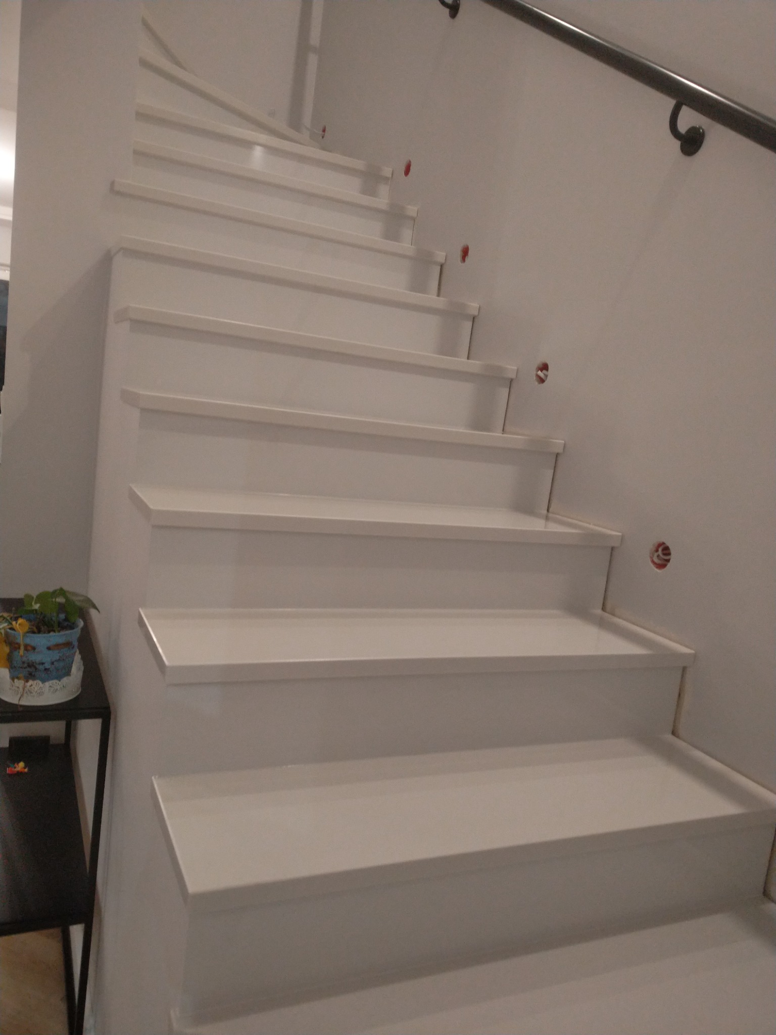 Białe schody z metalową poręczą, widoczne niedoskonałości w postaci otworów w ścianie i doniczka z kwiatami na stoliku obok.