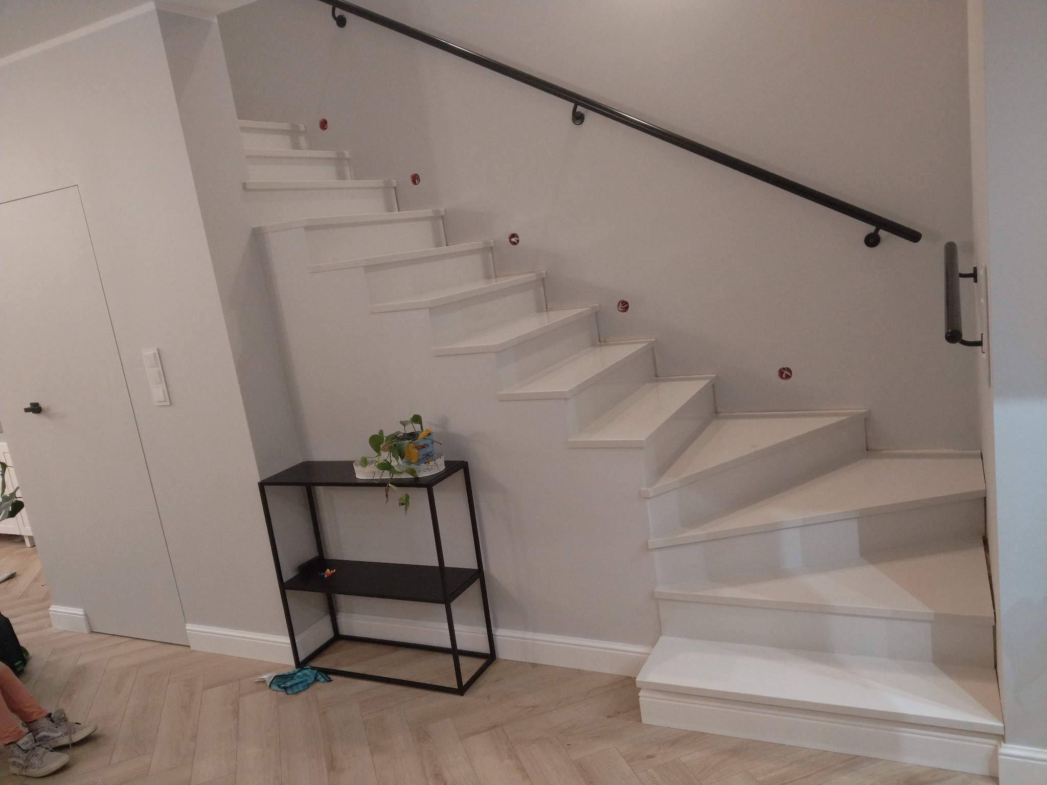 Białe, nowoczesne schody z czarną poręczą, widok z dołu, na podłodze panele imitujące drewno, obok schodów czarny regał z rośliną.