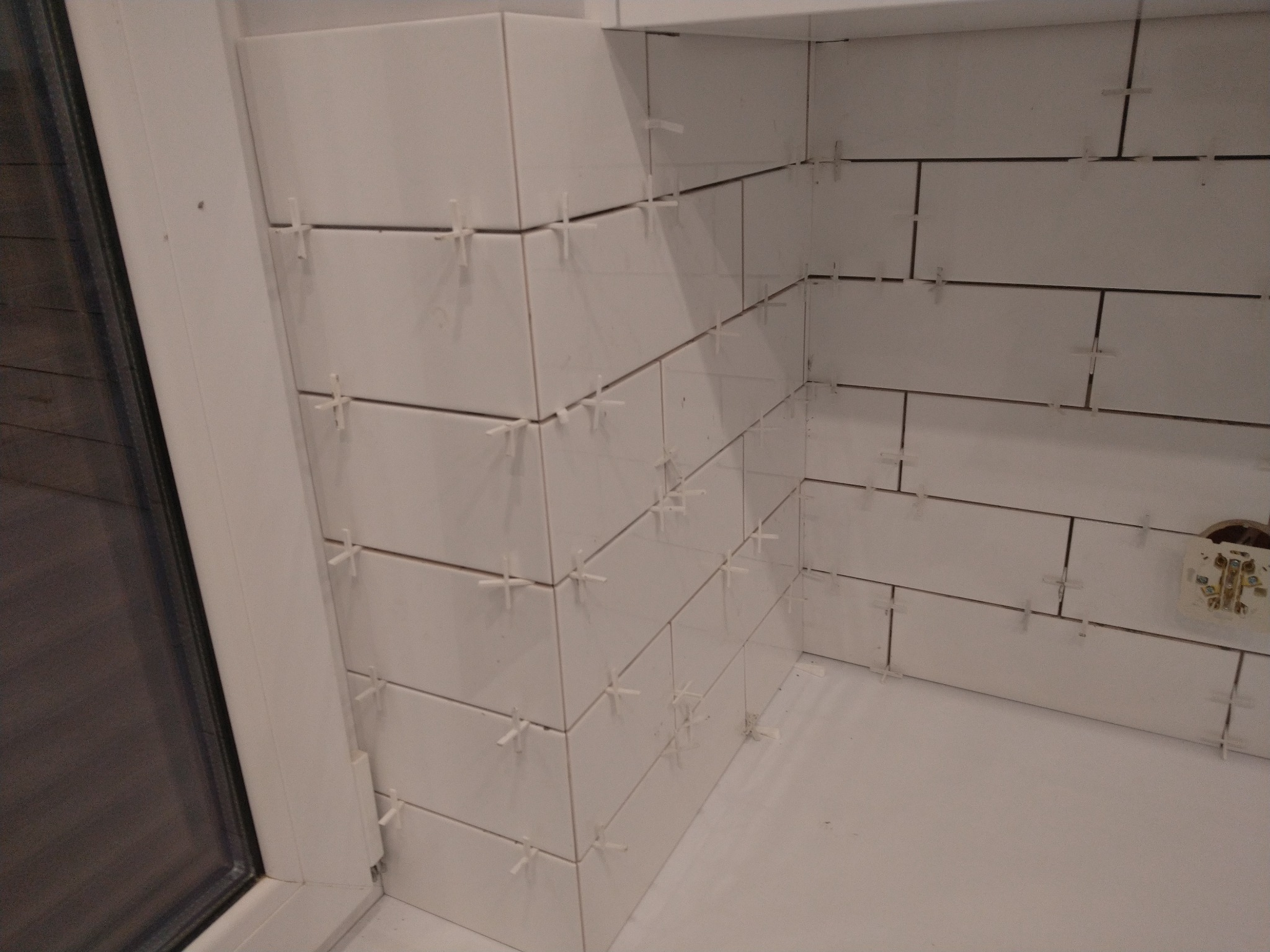 Białe, prostokątne płytki ułożone na ścianie i podłodze w rogu pomieszczenia, z plastikowymi krzyżykami dystansowymi w fugach, widoczna puszka elektryczna na ścianie.