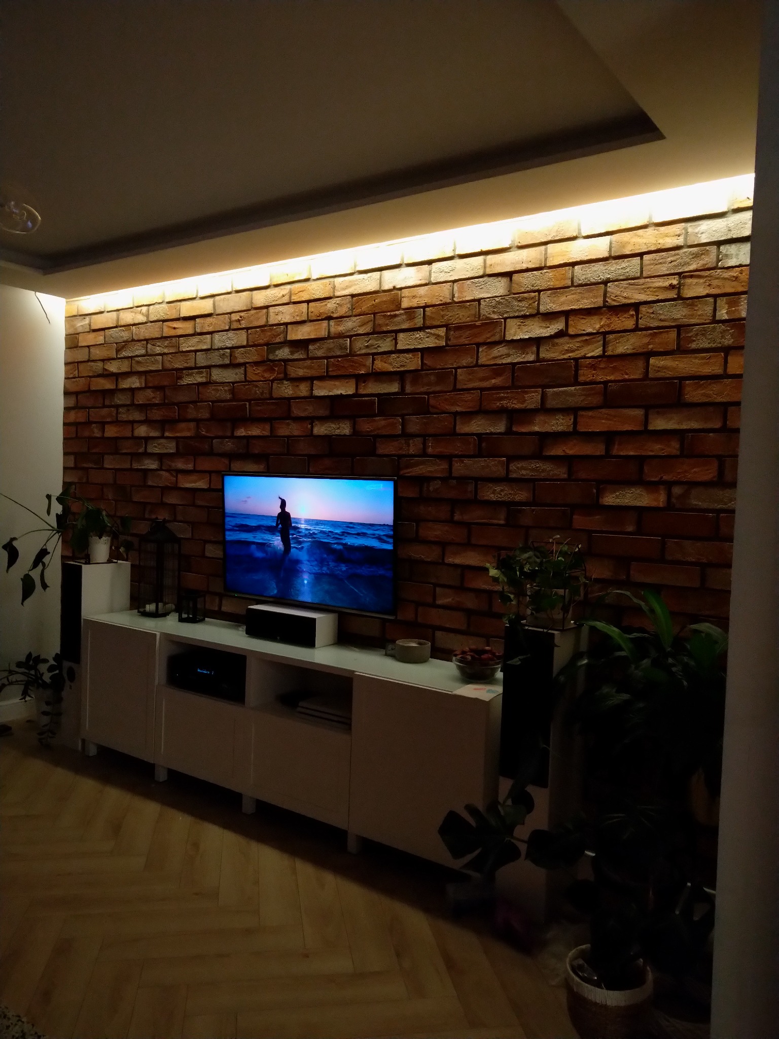 Salon z ceglaną ścianą podświetloną ledami, białą szafką RTV, telewizorem z widokiem na morze i parkietem w jodełkę.