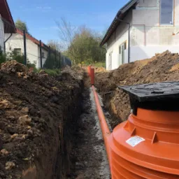 Pomarańczowa rura kanalizacyjna ułożona w wykopie ziemnym, widoczny zbiornik retencyjny z czarną pokrywą, w tle dom w stanie surowym i ogrodzenie sąsiada.