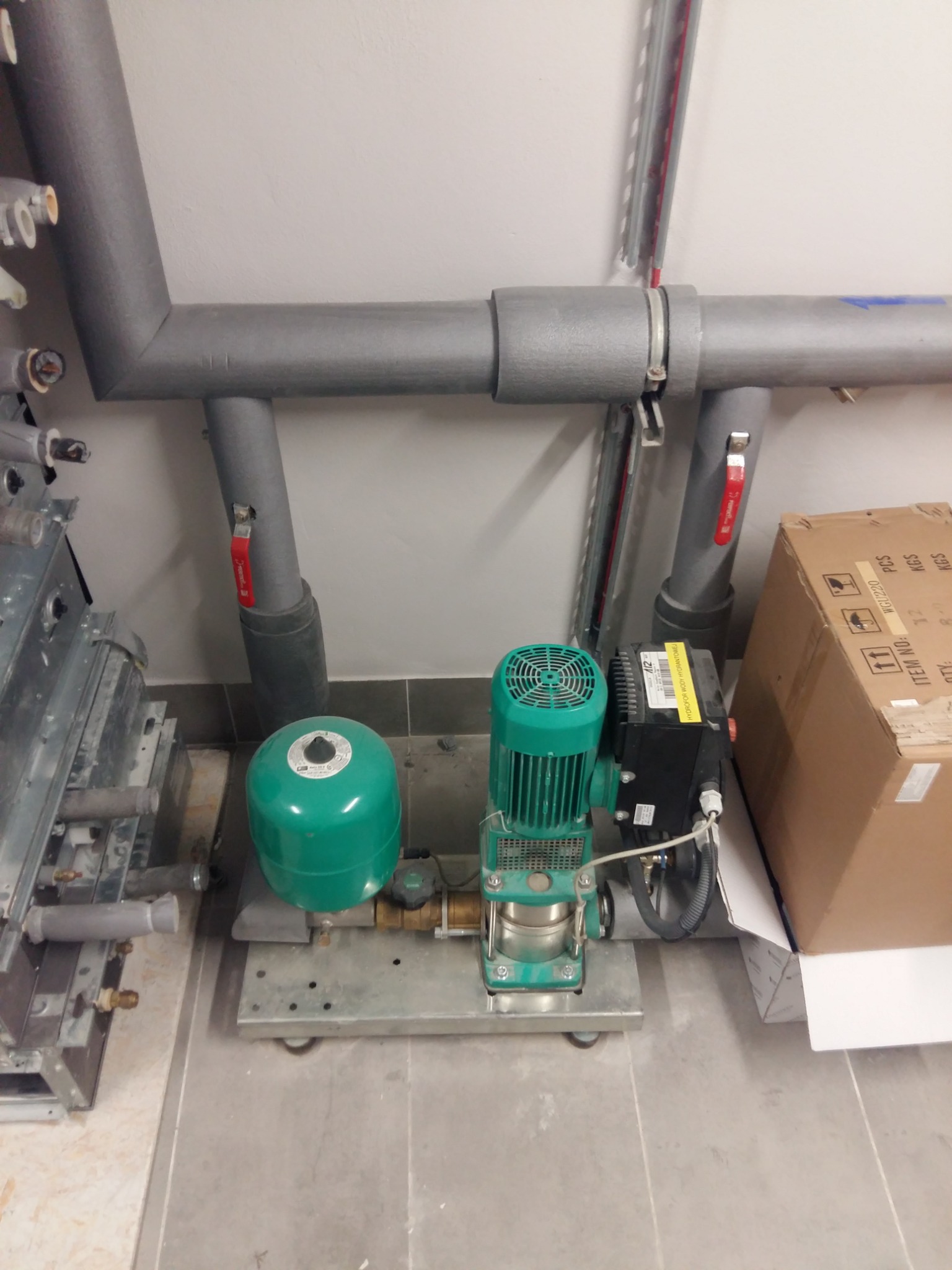 Zainstalowany hydrofor z zielonym zbiornikiem ciśnieniowym i pompą, widoczne szare rury instalacji wodnej z zaworami, zamocowane do ściany, obok kartonowe opakowanie