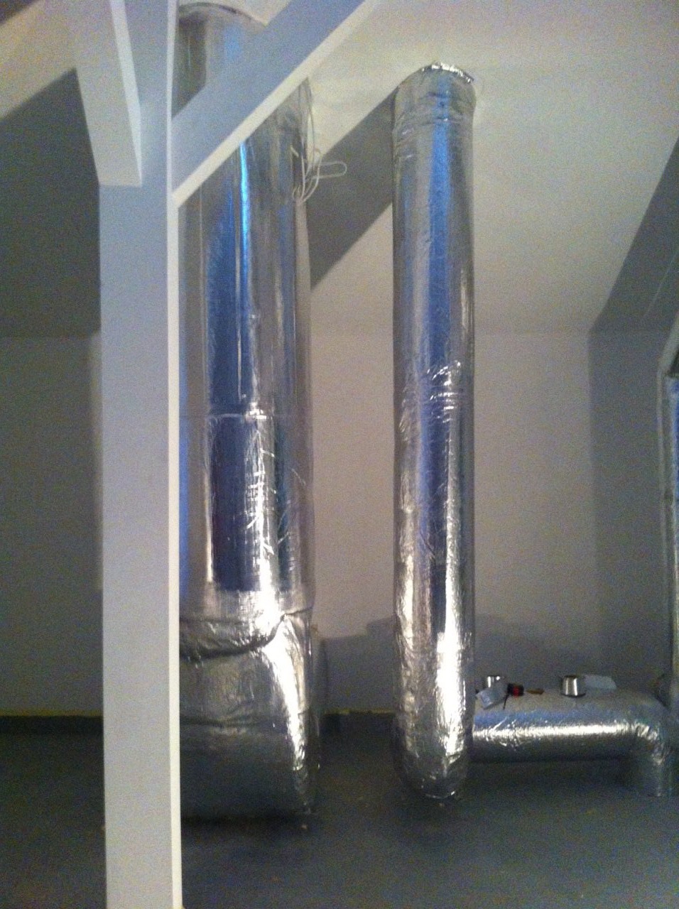 Dwie pionowe rury wentylacyjne owinięte folią aluminiową, z widocznym fragmentem konstrukcji drewnianej i fragmentem instalacji poziomej w tle.