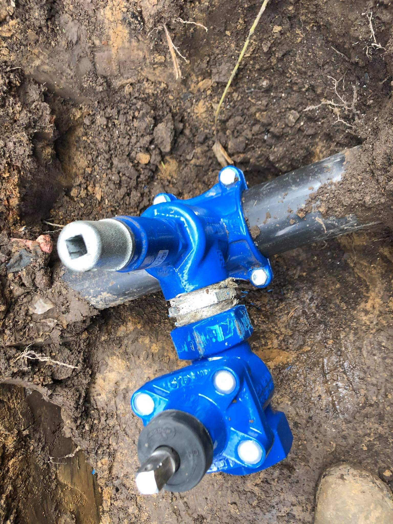 Niebieski zawór odcinający na czarnej rurze w wykopie ziemnym, widoczne połączenia śrubowe i nasadka z kwadratowym otworem.