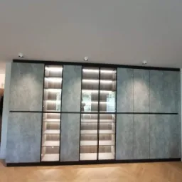 Duża, szara szafa wnękowa z podświetlanymi p&oacute;łkami za szklanymi drzwiami, wbudowana w ścianę na drewnianej podłodze ułożonej w jodełkę.