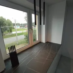 Czarny blat z marmurowym wzorem przy oknie, z widokiem na zewnątrz, zestaw noży w bloku, minimalistyczne lampy wiszące.