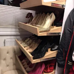 Wysuwane półki w garderobie prezentują kolekcję obuwia, od szpilek z brokatem po sportowe sneakersy, z bliska widoczne detale mechanizmów wysuwu i tapicerowany fotel obok.