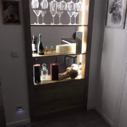 Drewniana witryna z oświetleniem LED, na półkach szkło koktajlowe, kieliszki do wina, butelki alkoholi oraz ozdobne pudełka, na ścianie obrazy.