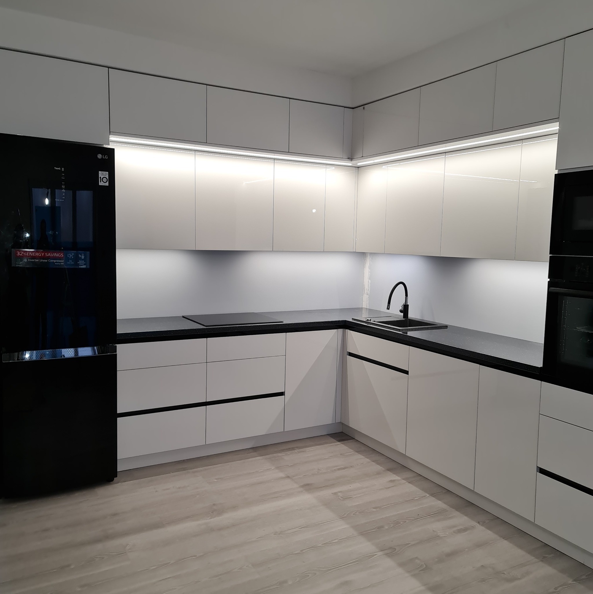 Nowoczesna, minimalistyczna kuchnia w Płońsku: białe, lakierowane fronty szafek z czarnymi, poziomymi uchwytami, czarny blat i sprzęt AGD, oświetlenie LED pod szafkami wiszącymi.