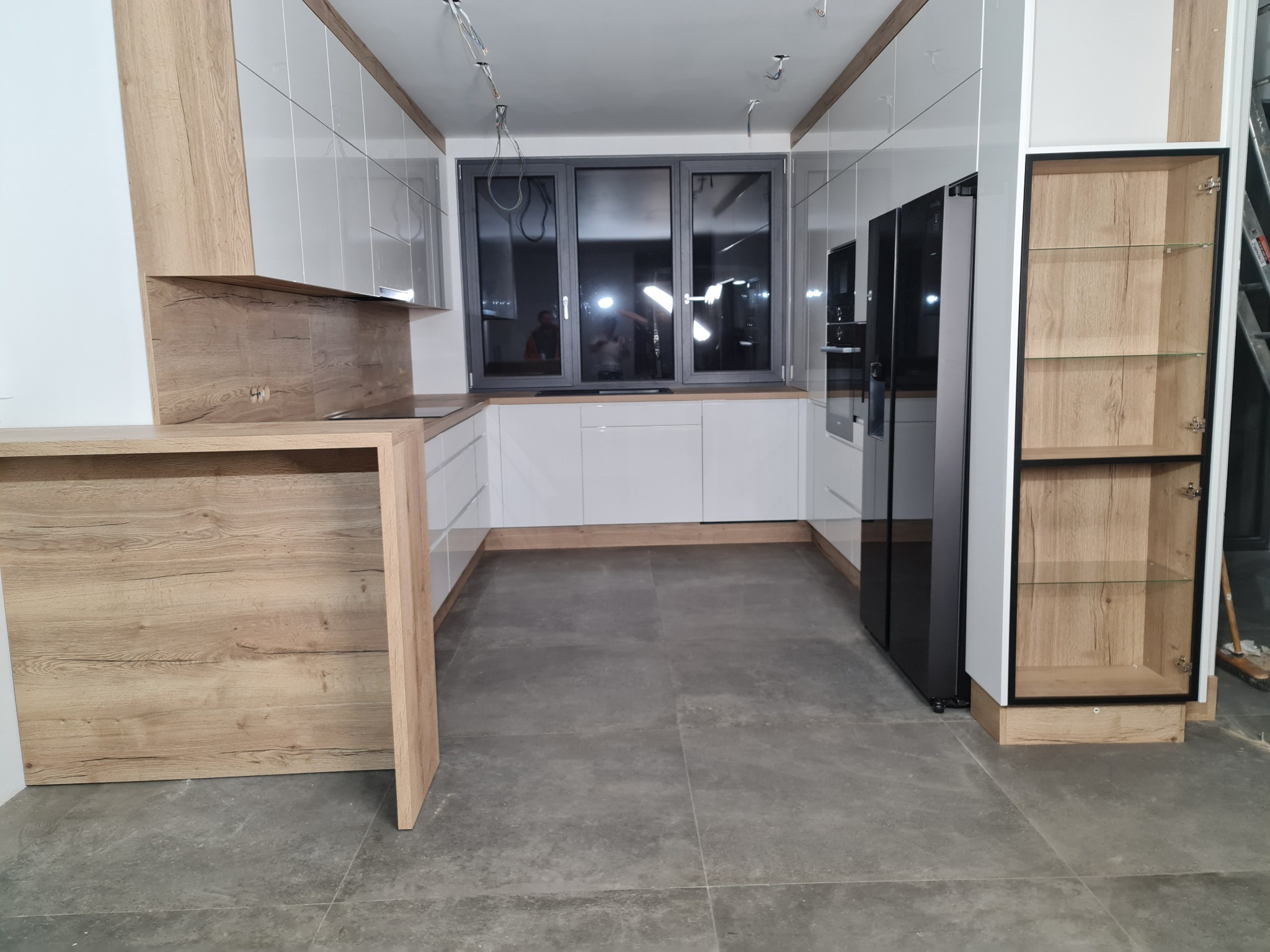 Nowoczesna kuchnia z białymi, lakierowanymi frontami szafek i drewnianym blatem, widoczne okno z trzema skrzydłami i czarna lodówka side-by-side, surowa podłoga z szarych płytek.