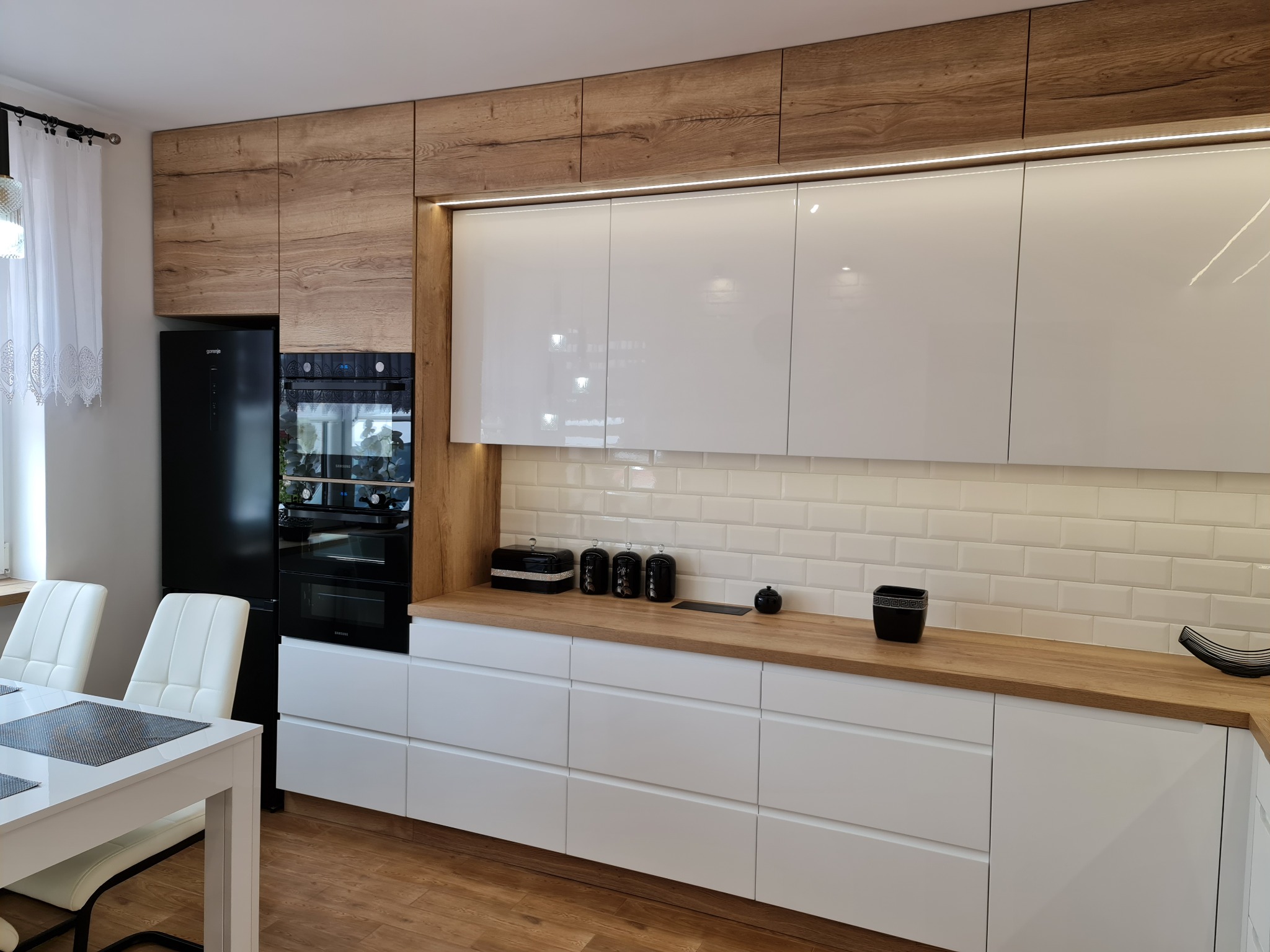 Nowoczesna kuchnia z białymi, lakierowanymi frontami szafek dolnych i górnych, drewnopodobnymi panelami górnymi, blatem drewnianym i białymi płytkami typu metro na ścianie, oświetlona listwą LED...