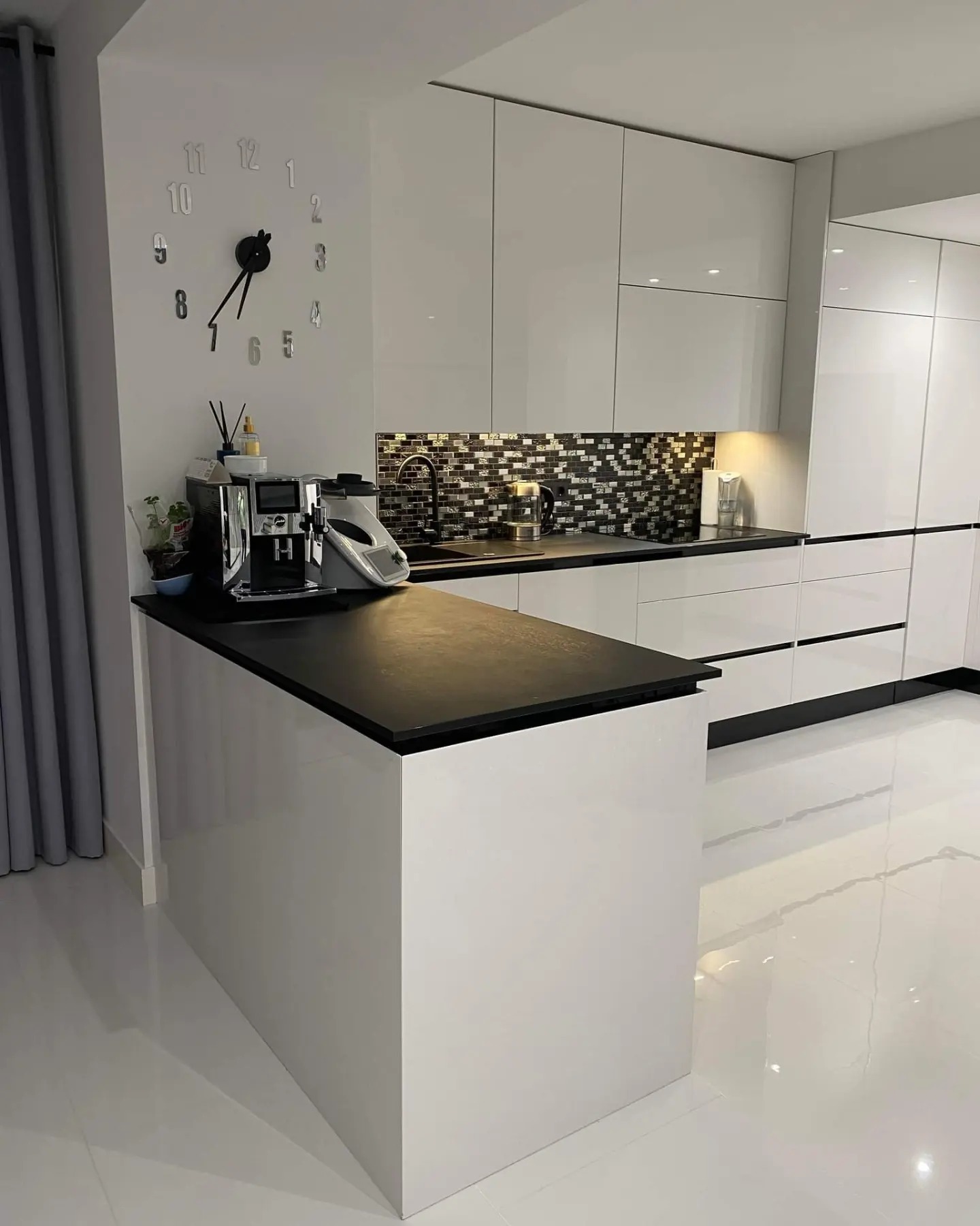 Nowoczesna, biała kuchnia z czarnym blatem i mozaiką nad blatem, uzupełniona o ekspres do kawy i designerski zegar ścienny.