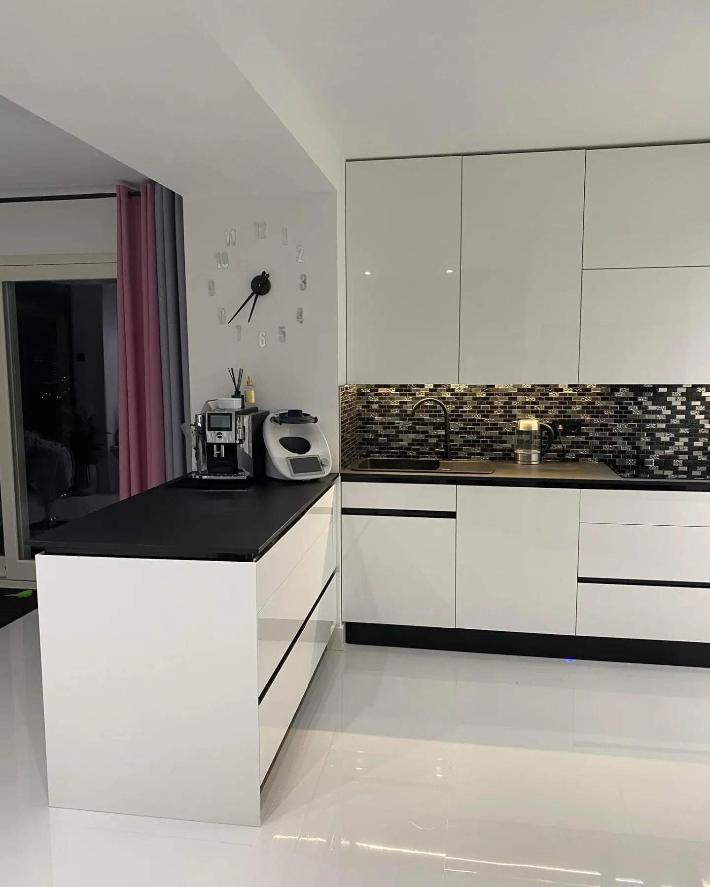 Nowoczesna kuchnia z białymi, lakierowanymi frontami, czarnym blatem i mozaiką nad zlewem. Na wyspie kuchennej ekspres do kawy i robot kuchenny.