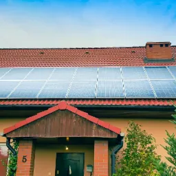 Dom jednorodzinny z czerwoną dachówką i panelami słonecznymi na dachu, widok z przodu, numer domu '5' widoczny na ścianie.