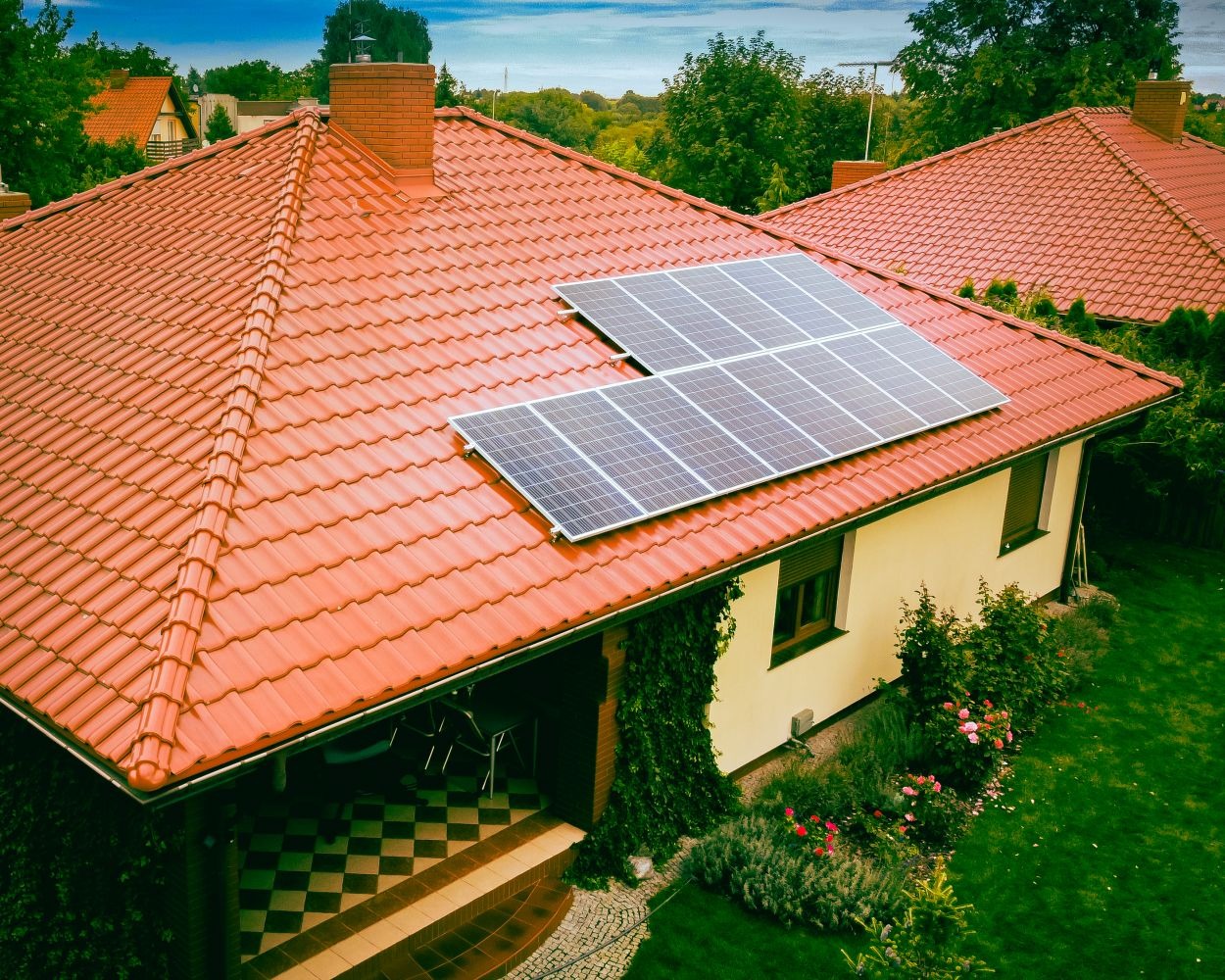 Widok z góry na dom z czerwoną dachówką, na której zamontowano dwa panele fotowoltaiczne. Ogród z kwiatami i zielenią wokół domu.