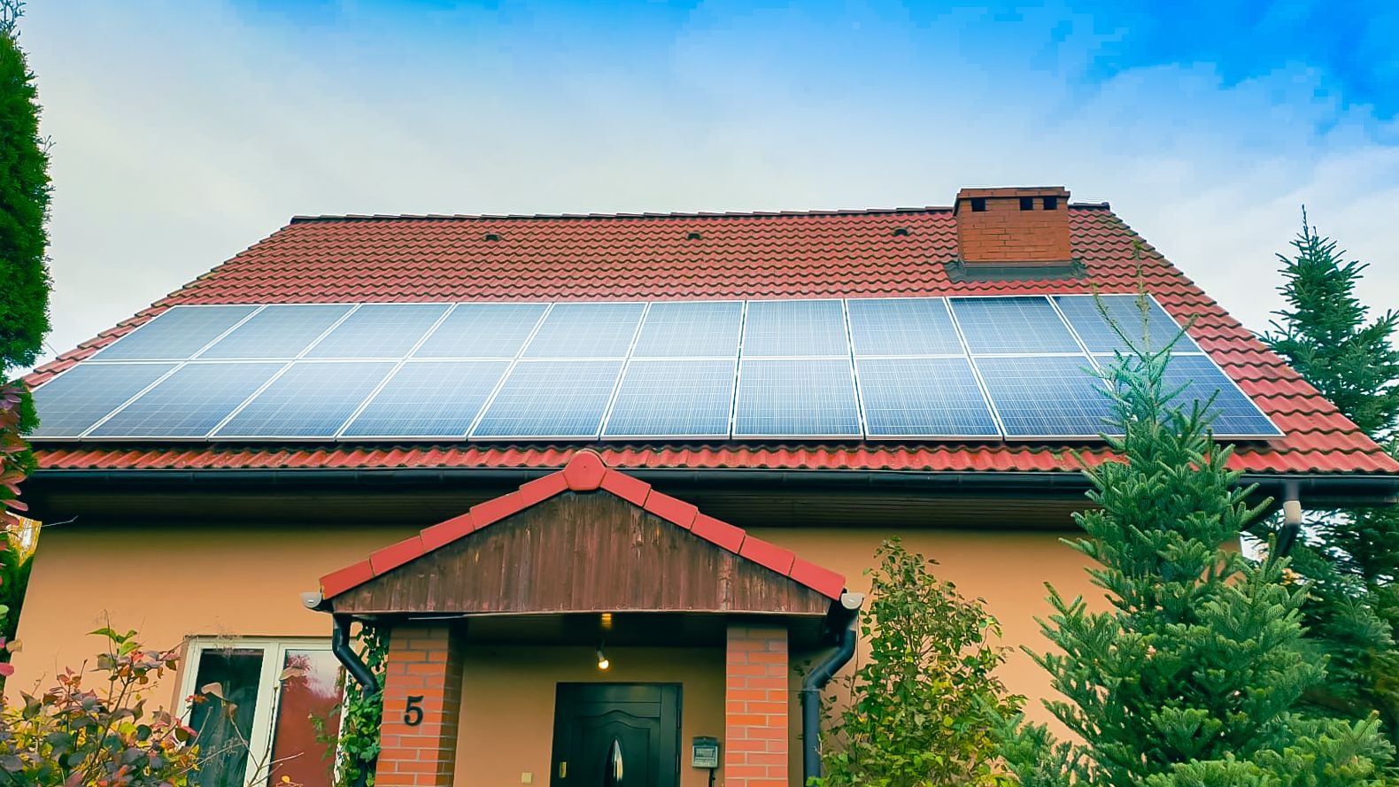Dom jednorodzinny z czerwoną dachówką i panelami słonecznymi na dachu, widok z przodu, numer domu '5' widoczny na ścianie.