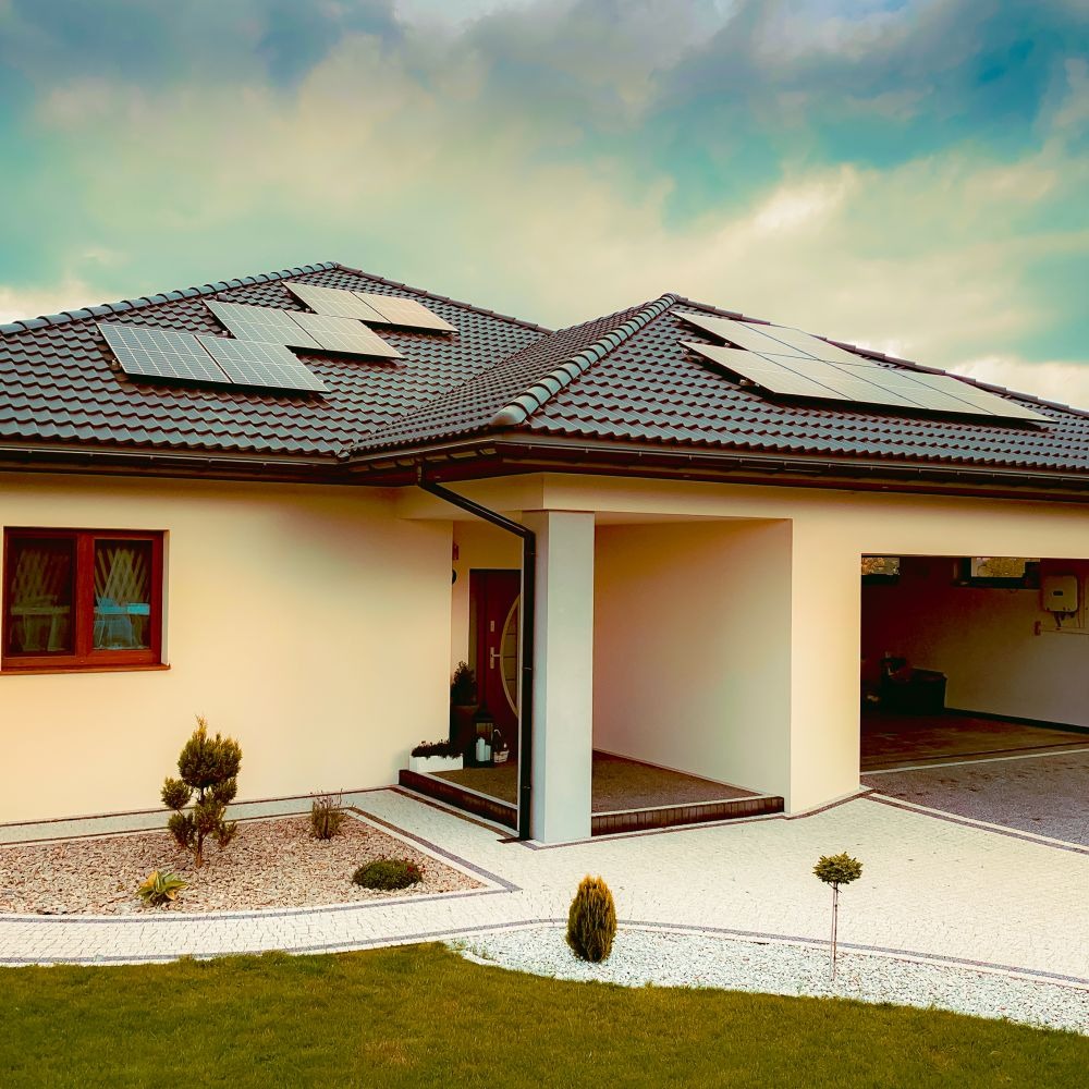 Dom jednorodzinny z zamontowanymi panelami słonecznymi na dachu, widok z przodu, jasna elewacja, brązowa dachówka, ozdobny ogródek z kamykami i niskimi krzewami.