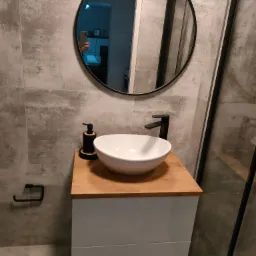 Nowoczesna łazienka z okrągłym lustrem, umywalką nablatową na drewnianym blacie i szarymi płytkami imitującymi beton.