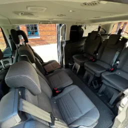 Wnętrze przestronnego busa z trzema rzędami siedzeń, widoczne uchwyty na ubrania i otwarte drzwi przesuwne, w tle ceglany budynek.