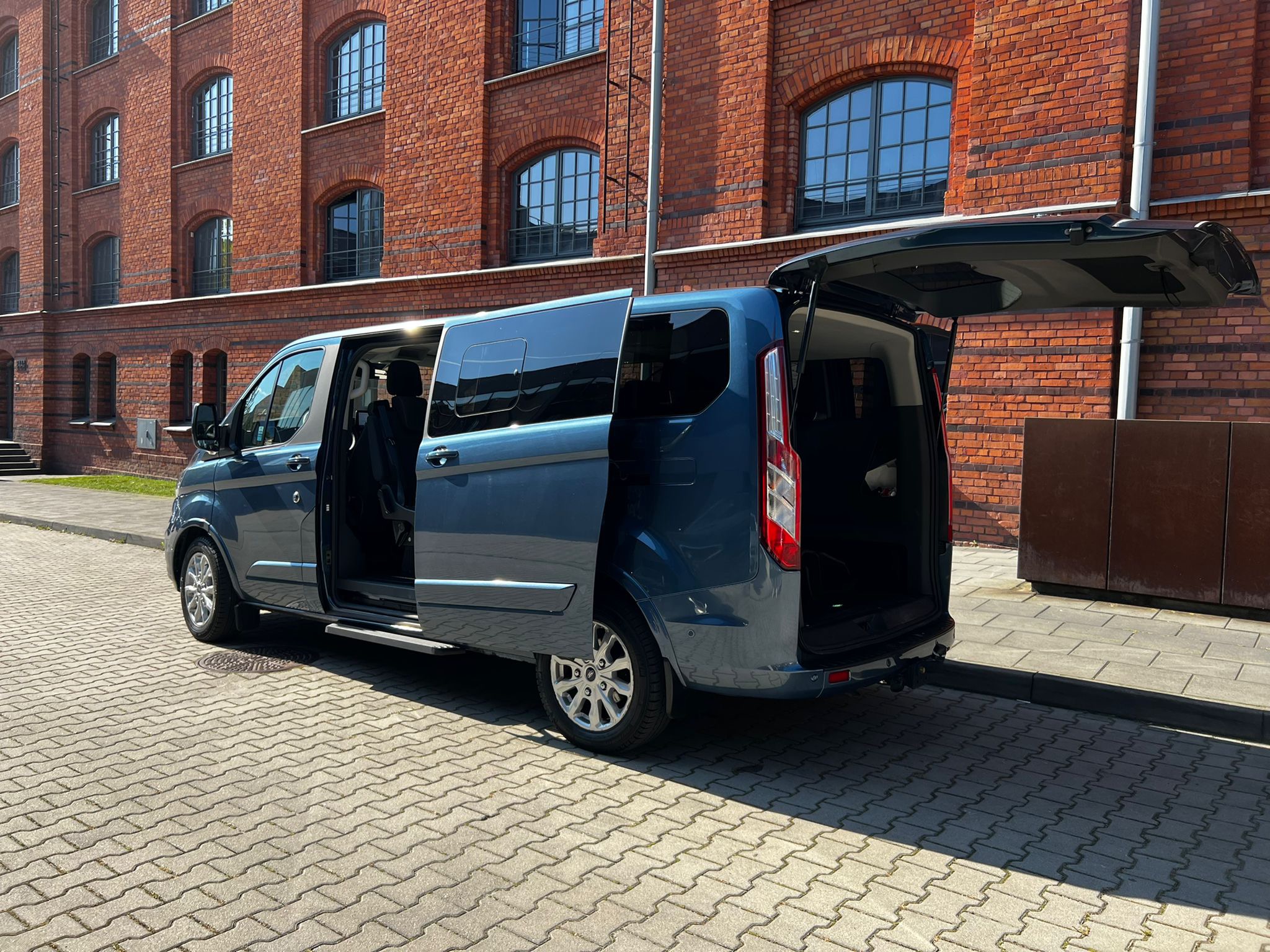 Niebieski van z otwartymi drzwiami i klapą bagażnika, zaparkowany na brukowanej ulicy przed budynkiem z czerwonej cegły.
