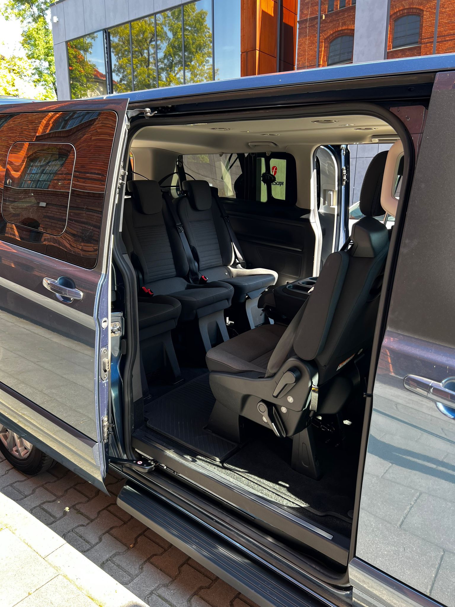 Wnętrze szarego vana z otwartymi drzwiami przesuwnymi, widoczne trzy rzędy foteli z czarną tapicerką i regulowanymi zagłówkami, logo firmy widoczne na szybie, budynek z cegły odbija się w szybach.