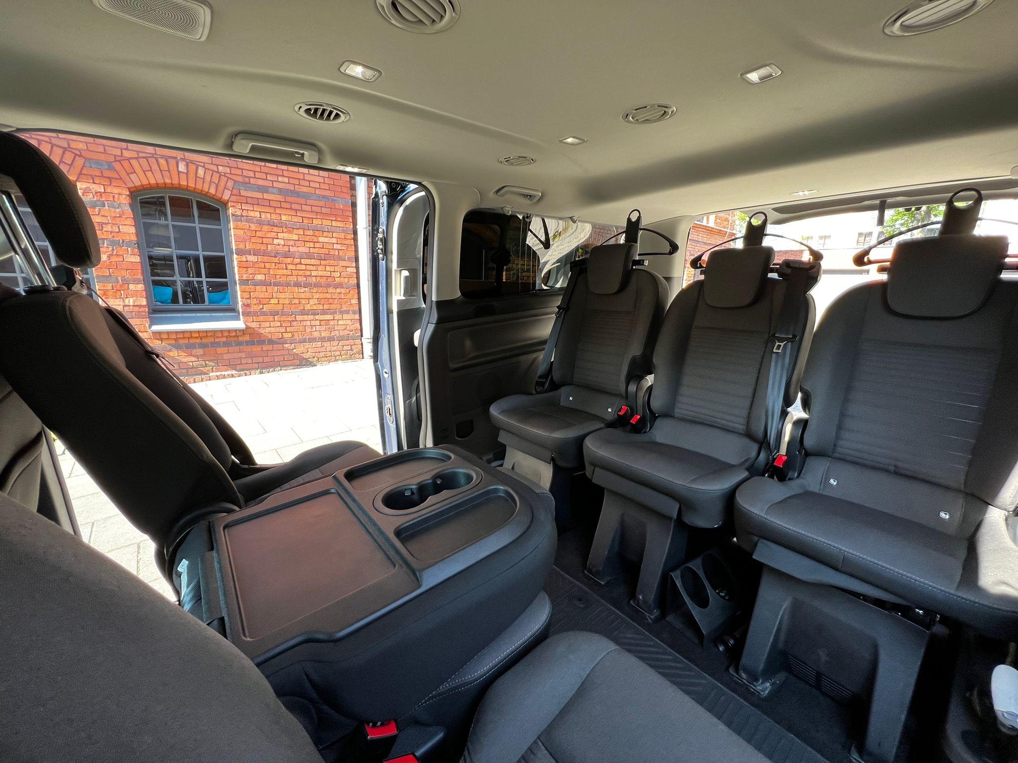 Wnętrze busa pasażerskiego z rozkładanym stolikiem, widok na fotele z wieszakami i ceglany budynek za oknem.