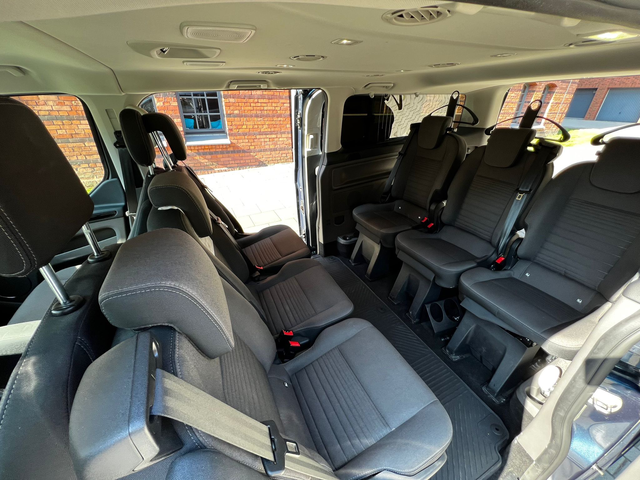 Wnętrze przestronnego busa z trzema rzędami siedzeń, widoczne uchwyty na ubrania i otwarte drzwi przesuwne, w tle ceglany budynek.