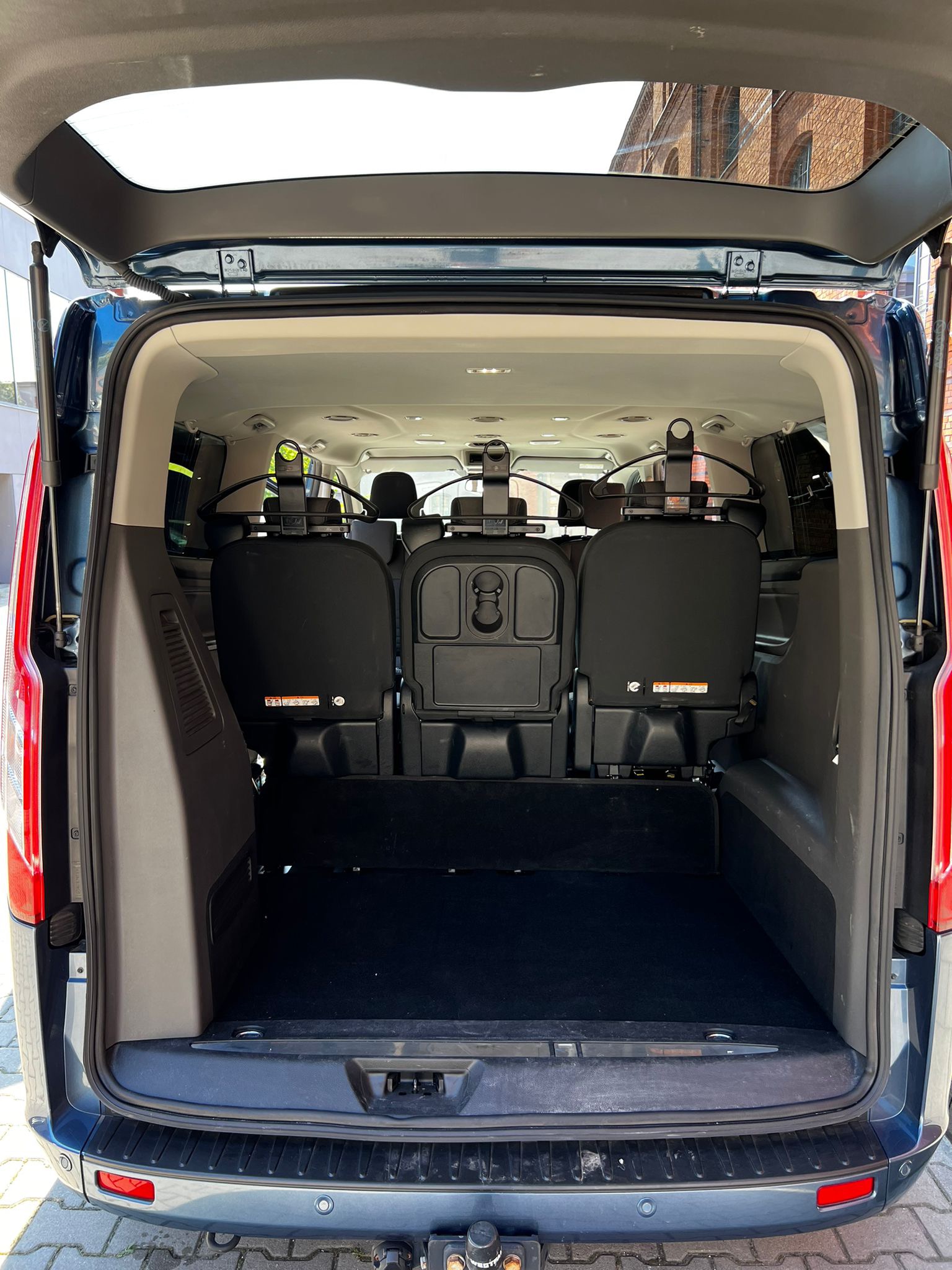 Wnętrze bagażnika busa z trzema rzędami siedzeń, z zamontowanymi wieszakami na ubrania na zagłówkach, widok z zewnątrz na otwartą klapę bagażnika.