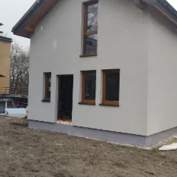 Nowoczesny dom jednorodzinny z szarą elewacją i drewnianymi oknami w trakcie wykańczania, widoczne niedokończone otoczenie i padający śnieg.