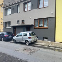 Everest bud - Dwukondygnacyjny budynek po renowacji elewacji, z szarą kolorystyką i drewnianymi oknami. Na tle błękitne niebo z chmurami. Na chodniku zaparkowane samochody.