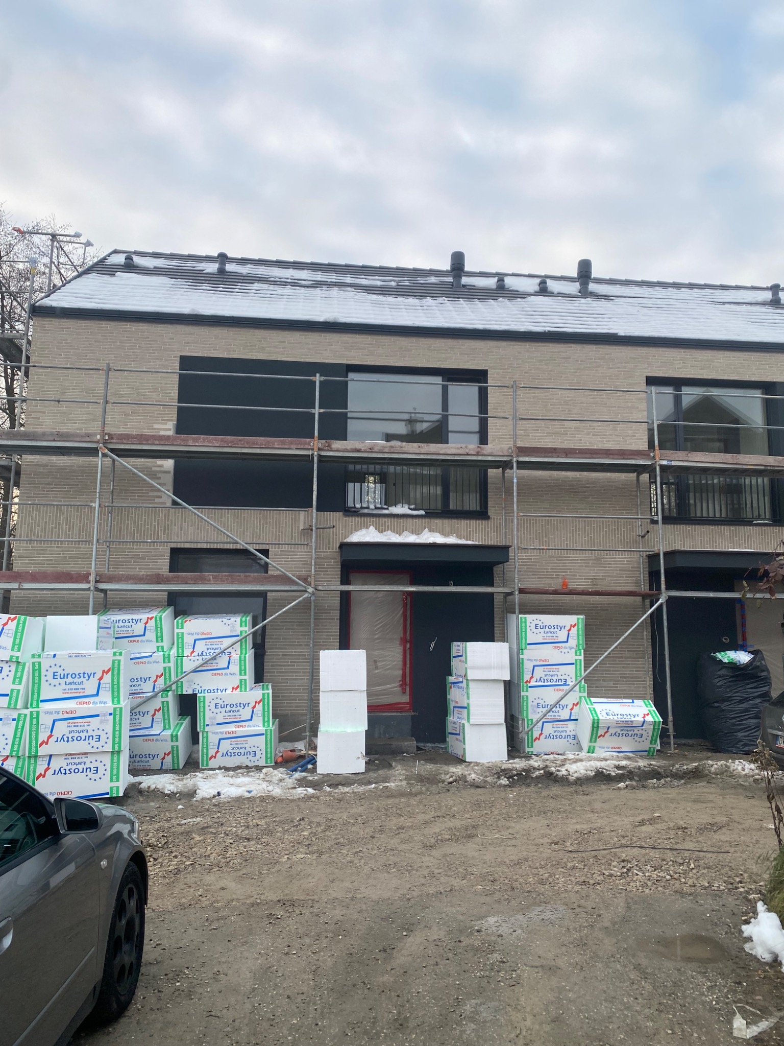 Ocieplanie elewacji domu z cegły. Na rusztowaniu, przed budynkiem, ułożone paczki styropianu Eurostyr. Widoczne okna z czarnymi ramami i fragment szarego samochodu.
