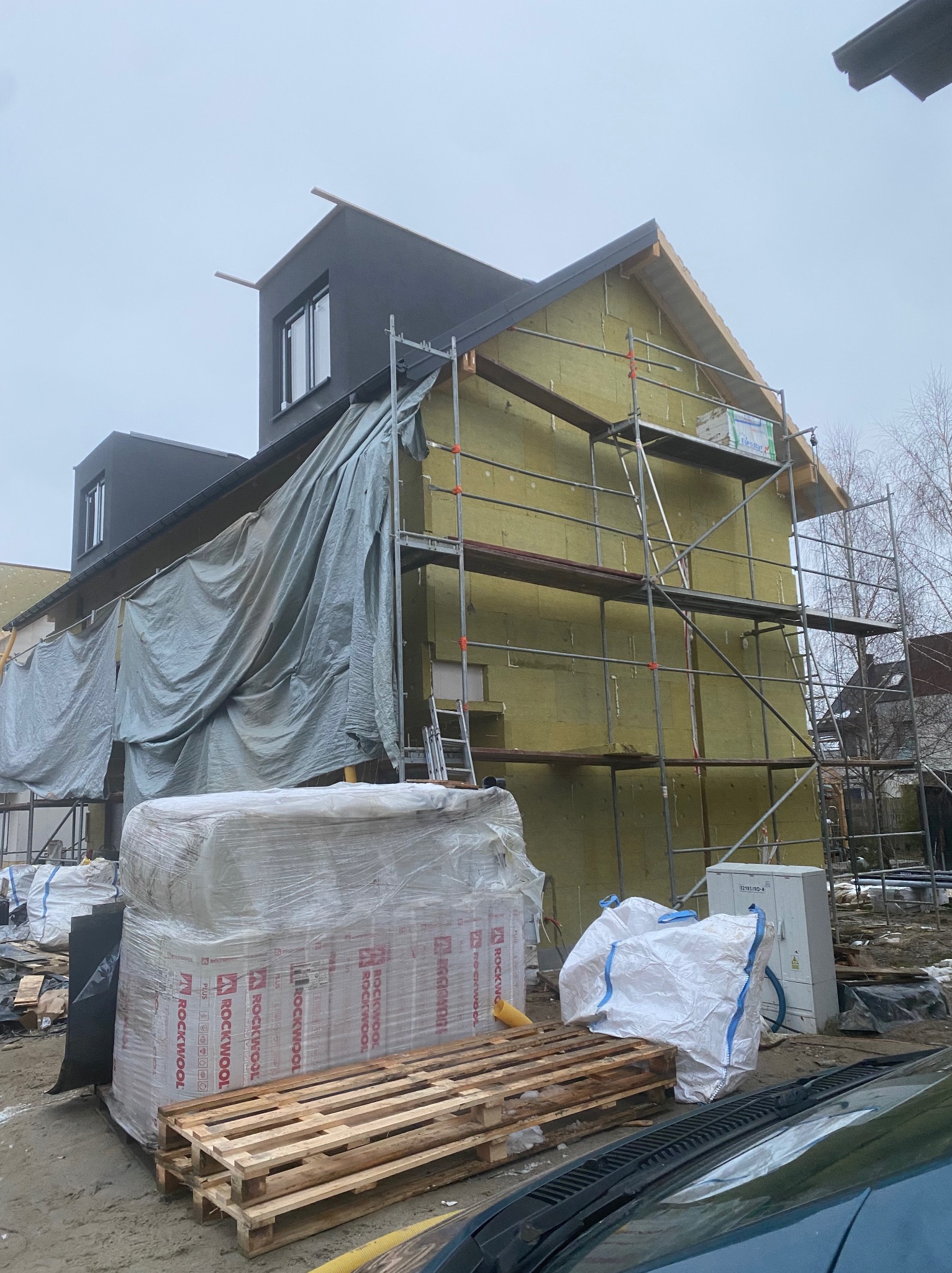 Ocieplanie domu wełną mineralną Rockwool, widoczne rusztowania i materiały budowlane. Część elewacji zakryta plandeką. Dom z czarną elewacją.