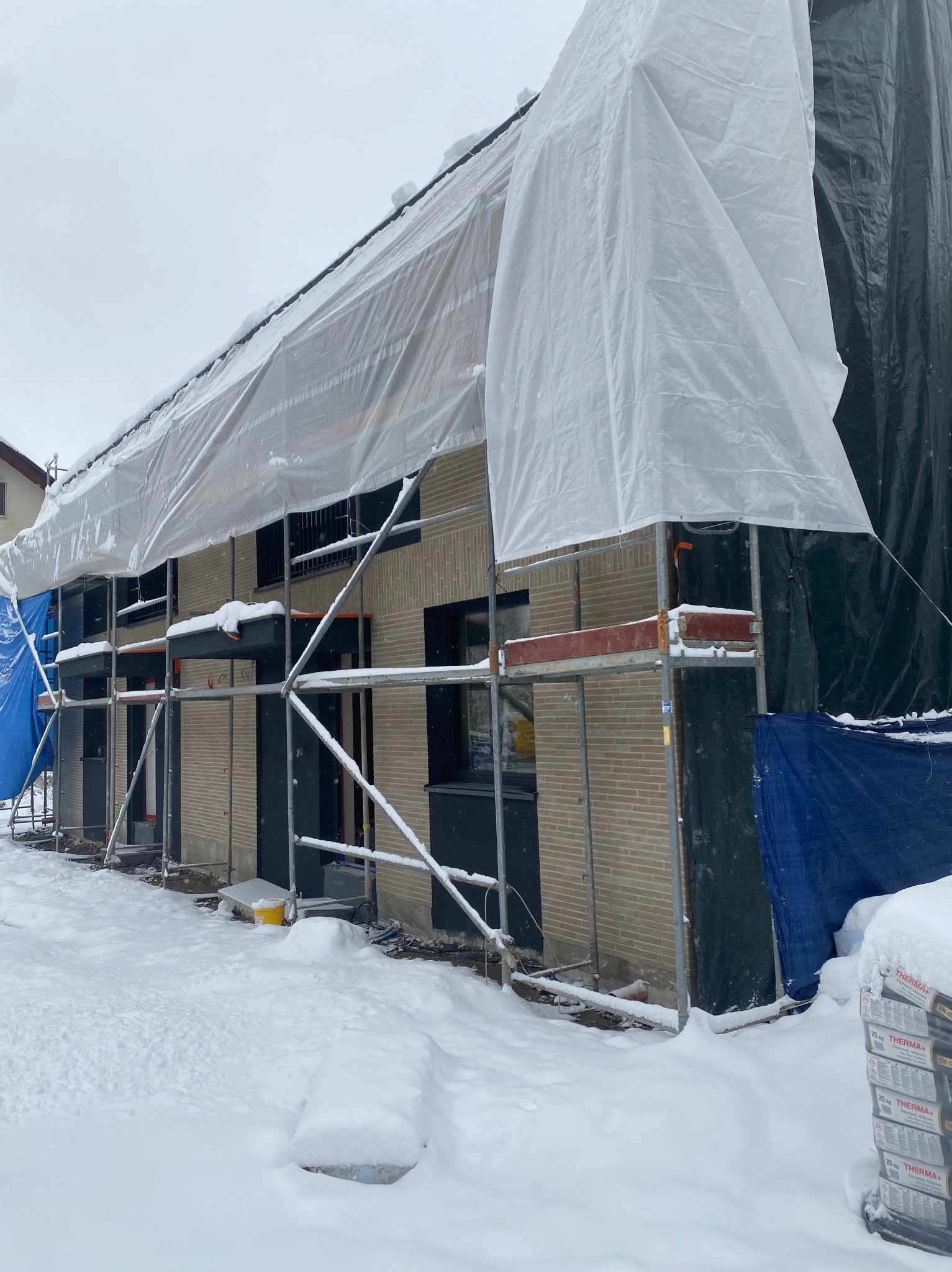 Dom w trakcie prac elewacyjnych zimą, osłonięty plandekami i rusztowaniami, z widocznymi paletami materiałów budowlanych. Śnieżny krajobraz.