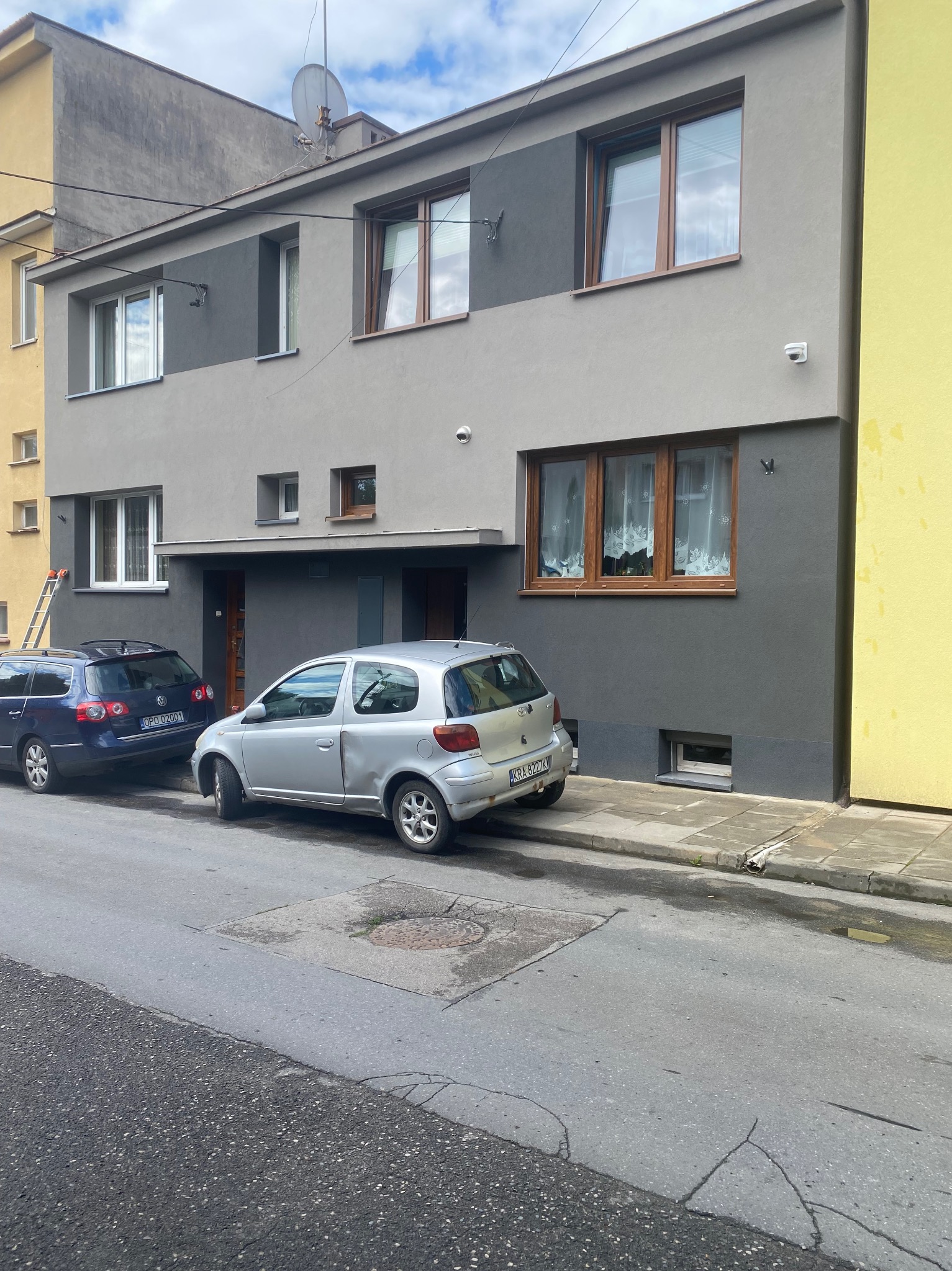 Dwukondygnacyjny budynek po renowacji elewacji, z szarą kolorystyką i drewnianymi oknami. Na tle błękitne niebo z chmurami. Na chodniku zaparkowane samochody.