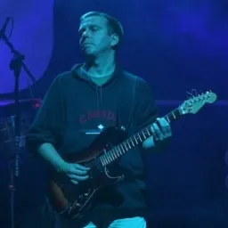 Mężczyzna w ciemnej bluzie z kapturem z napisem 'CANADA' gra na elektrycznej gitarze na scenie oświetlonej niebieskim światłem.