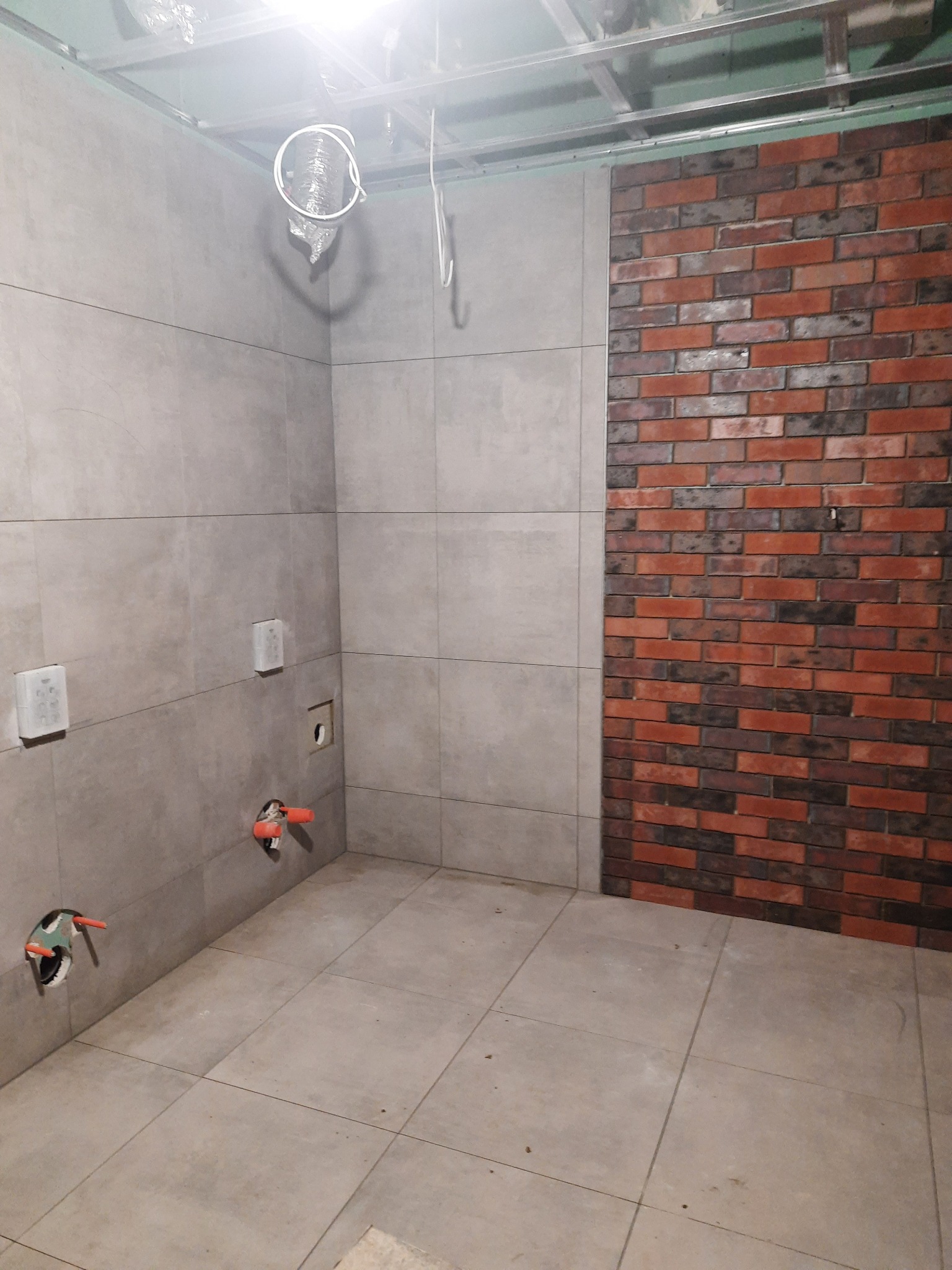 Wnętrze łazienki w trakcie wykańczania: szare płytki na ścianach i podłodze, fragment ściany wyłożony dekoracyjną czerwoną cegłą, wystające rury instalacji wodnej, widoczne elementy podwieszanego...